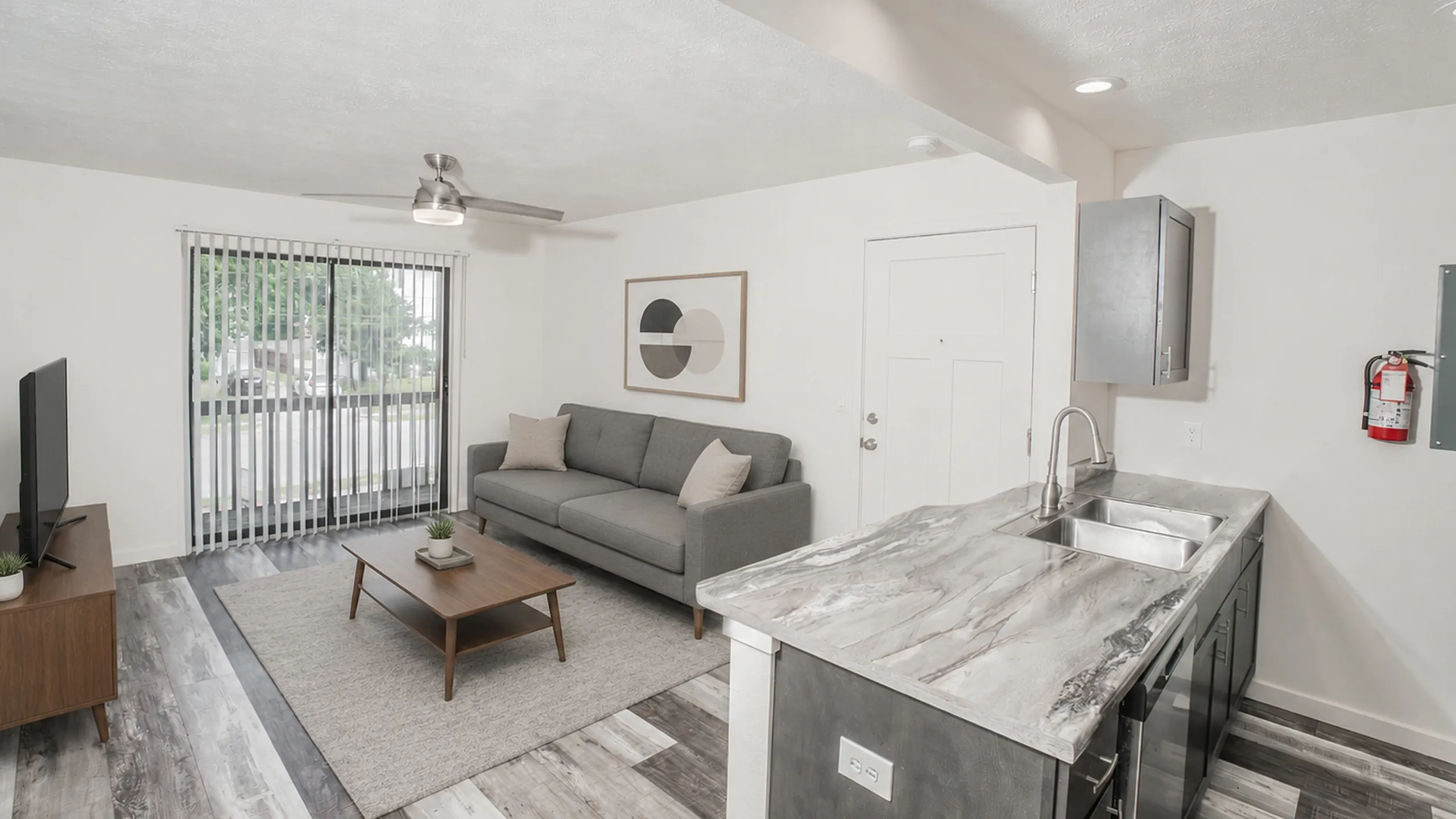Chic modern living area featuring a sleek gray sofa, stylish coffee table, large window with vertical blinds, and a sophisticated open-concept kitchen with elegant countertops.