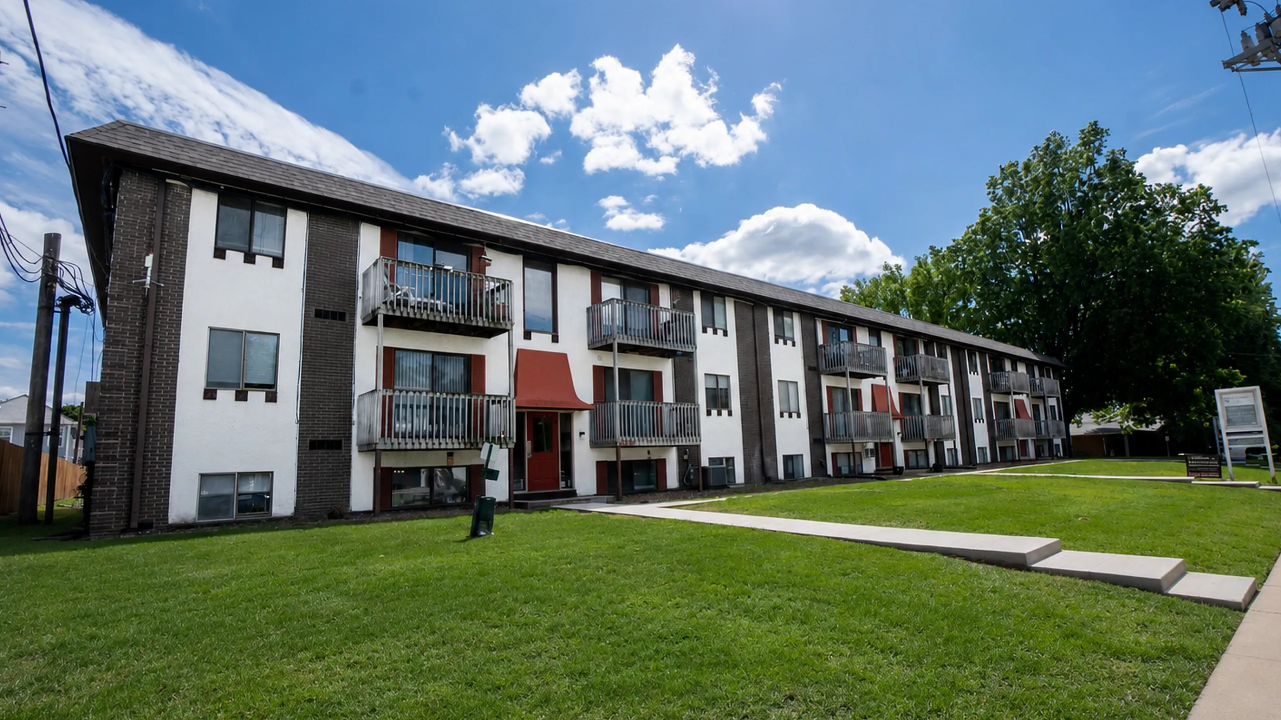 Charming three-story apartment building with private balconies, surrounded by lush green grass and under a bright blue sky.