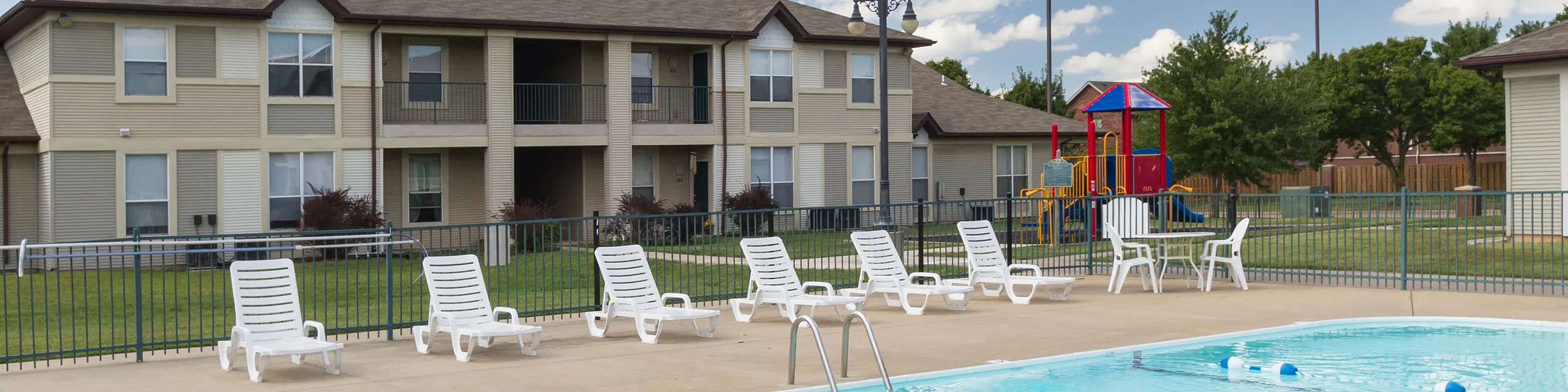 Charming apartment complex featuring a sparkling swimming pool, lounge chairs, and a colorful playground under a bright blue sky.