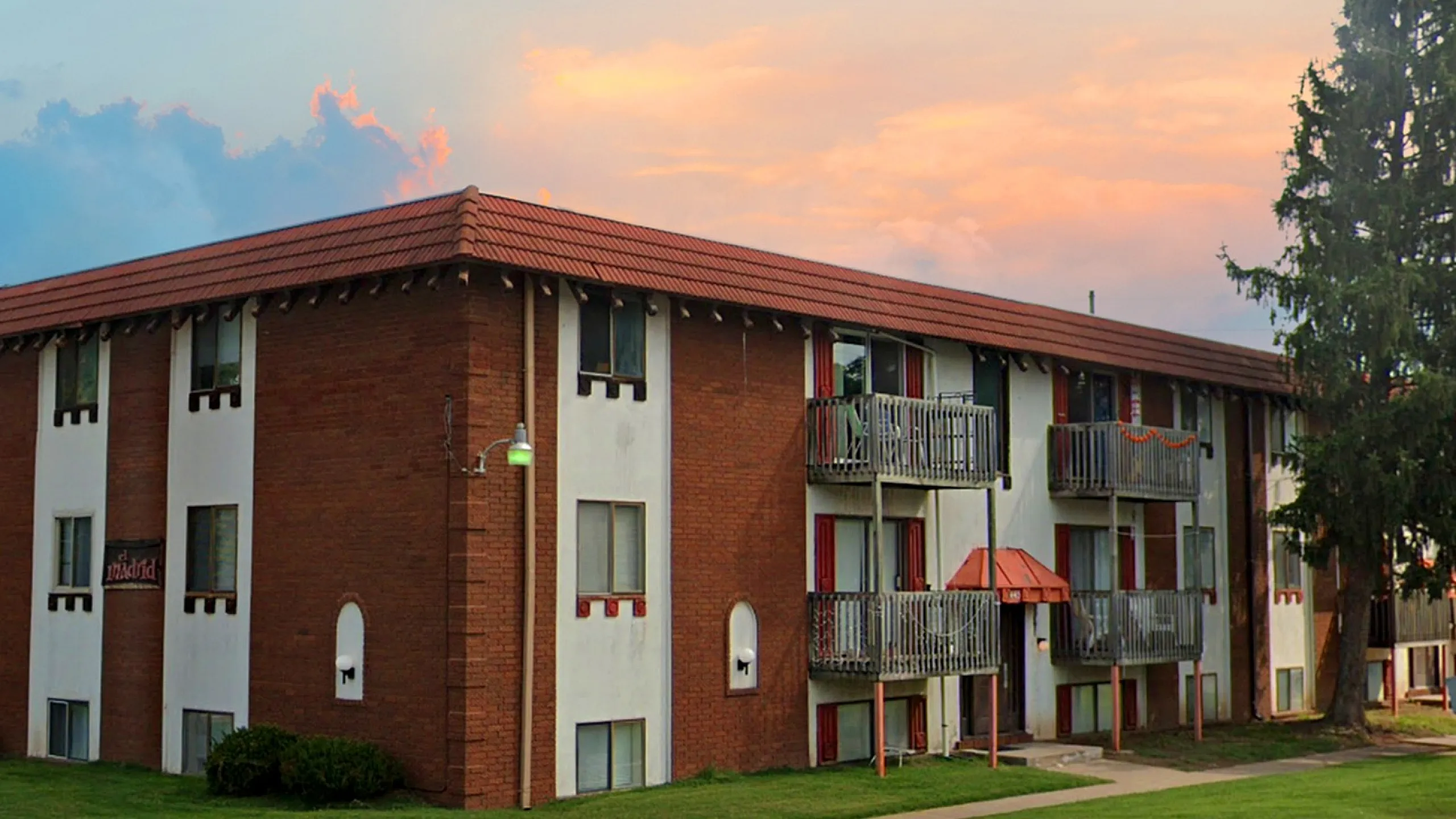 Charming brick apartment building with private balconies and lush surroundings, set against a picturesque sunset sky.