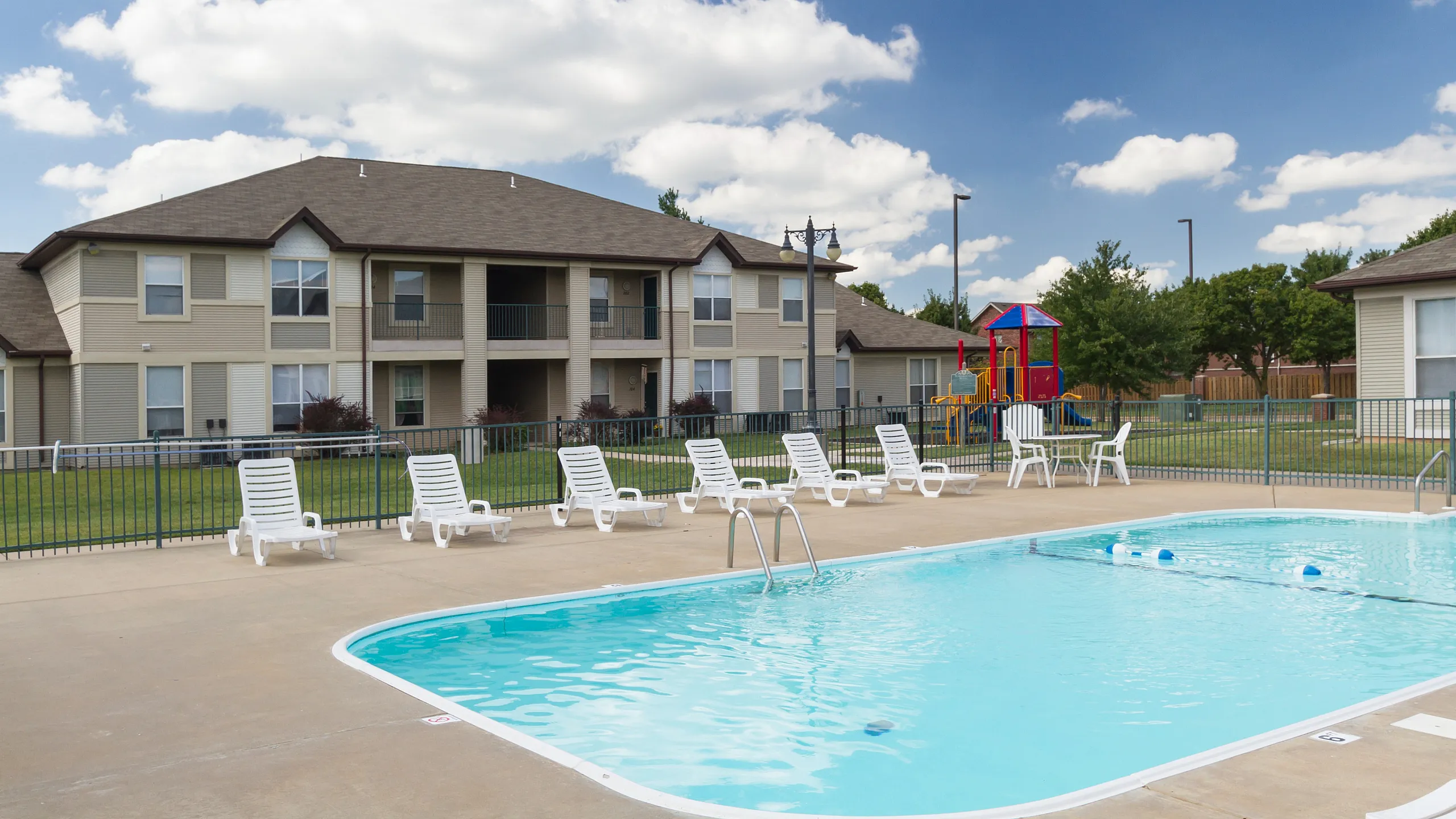 Charming apartment complex with bright blue swimming pool, sun loungers, and colorful playground under clear skies.