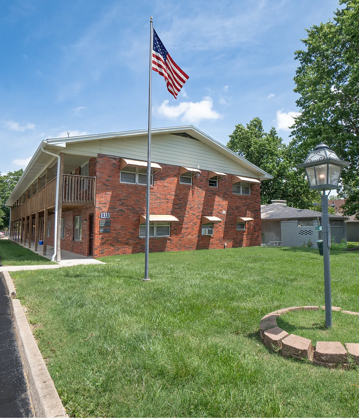 Charming brick apartment complex with lush green lawn and welcoming American flag, perfect for serene living.