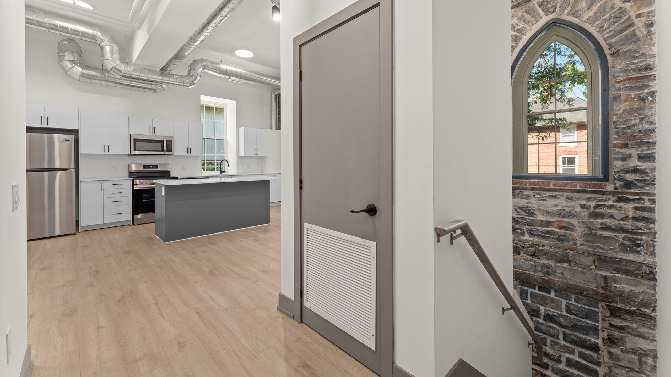 Modern kitchen with sleek white cabinetry, stainless steel appliances, and a large window, adjacent to a unique stone wall staircase.