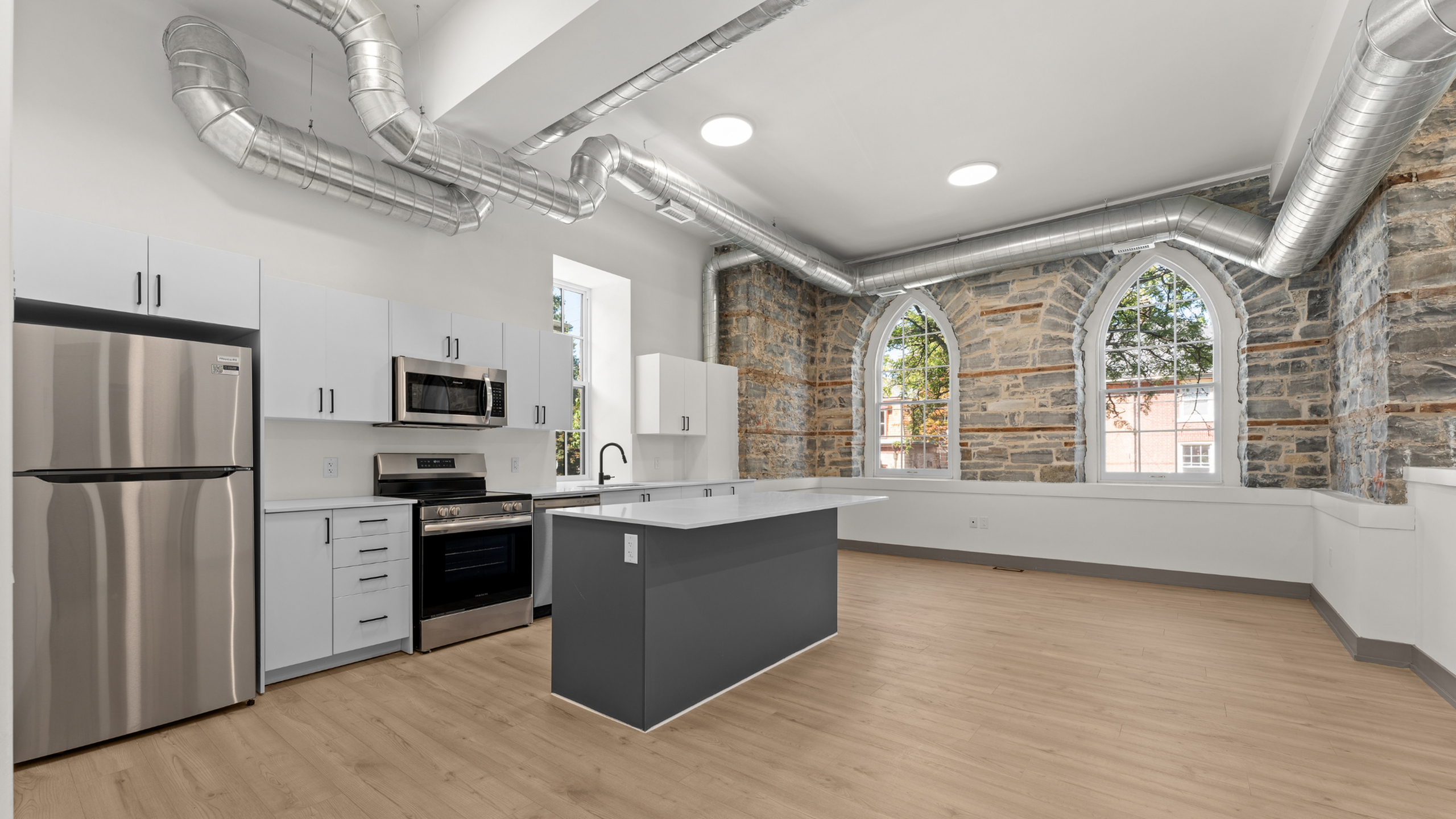 Modern kitchen with stainless steel appliances, exposed ductwork, and charming arched windows set against original stone walls.