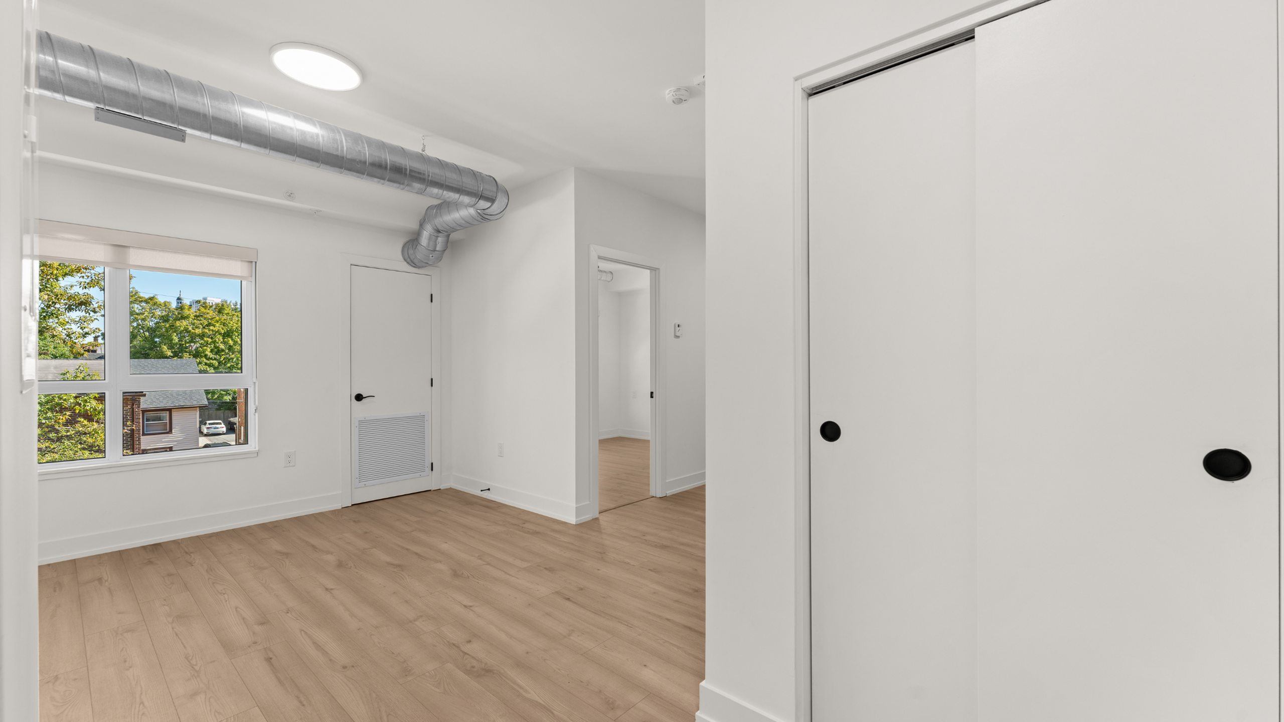 Bright and contemporary room with sleek hardwood flooring, large window with tree views, and modern exposed ductwork.