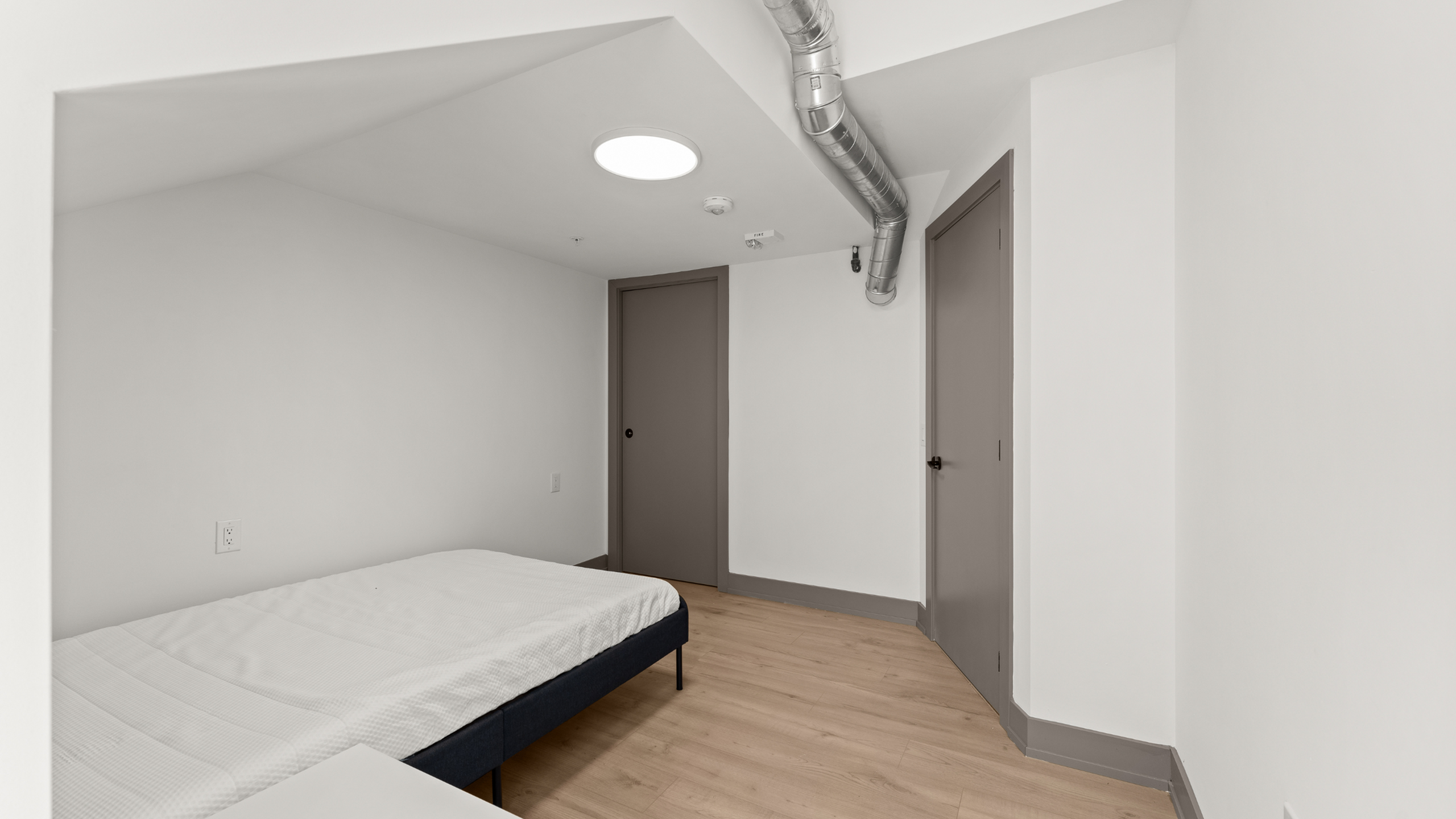 Modern minimalistic bedroom with clean white walls, cozy bed, and sleek wooden flooring, featuring stylish exposed ductwork and ample lighting.
