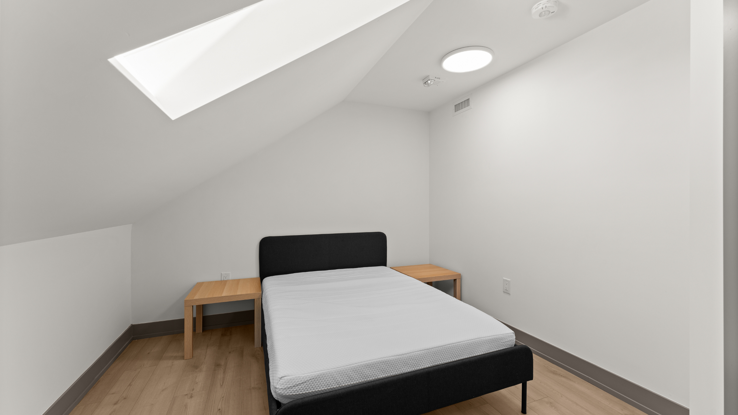 Modern minimalist bedroom with sloped ceiling, featuring a sleek bed and wooden side tables, illuminated by natural light from a skylight.