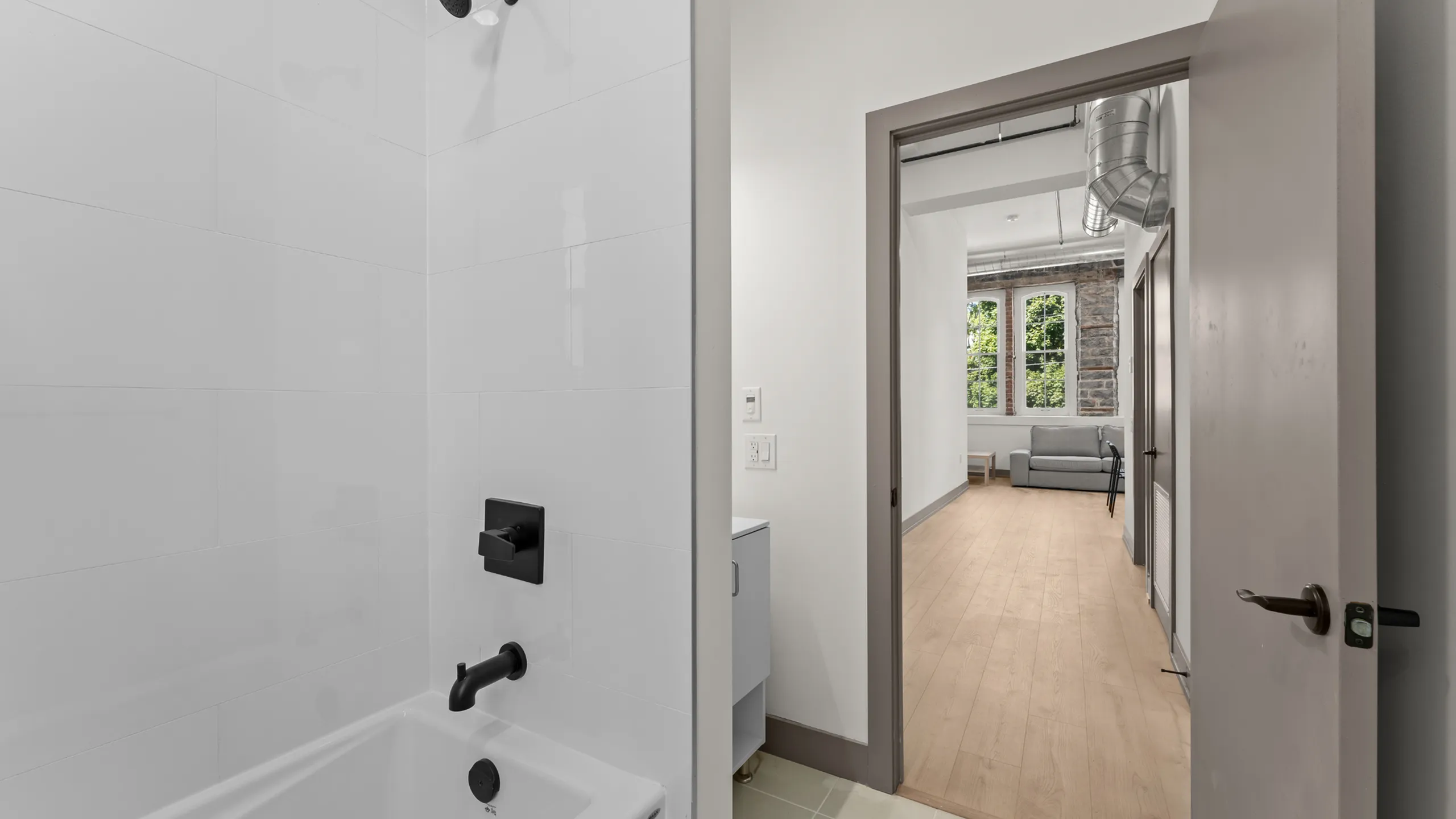 Modern apartment interior featuring a sleek white bathroom leading to a bright hallway with wooden floors and a cozy seating area by large windows.