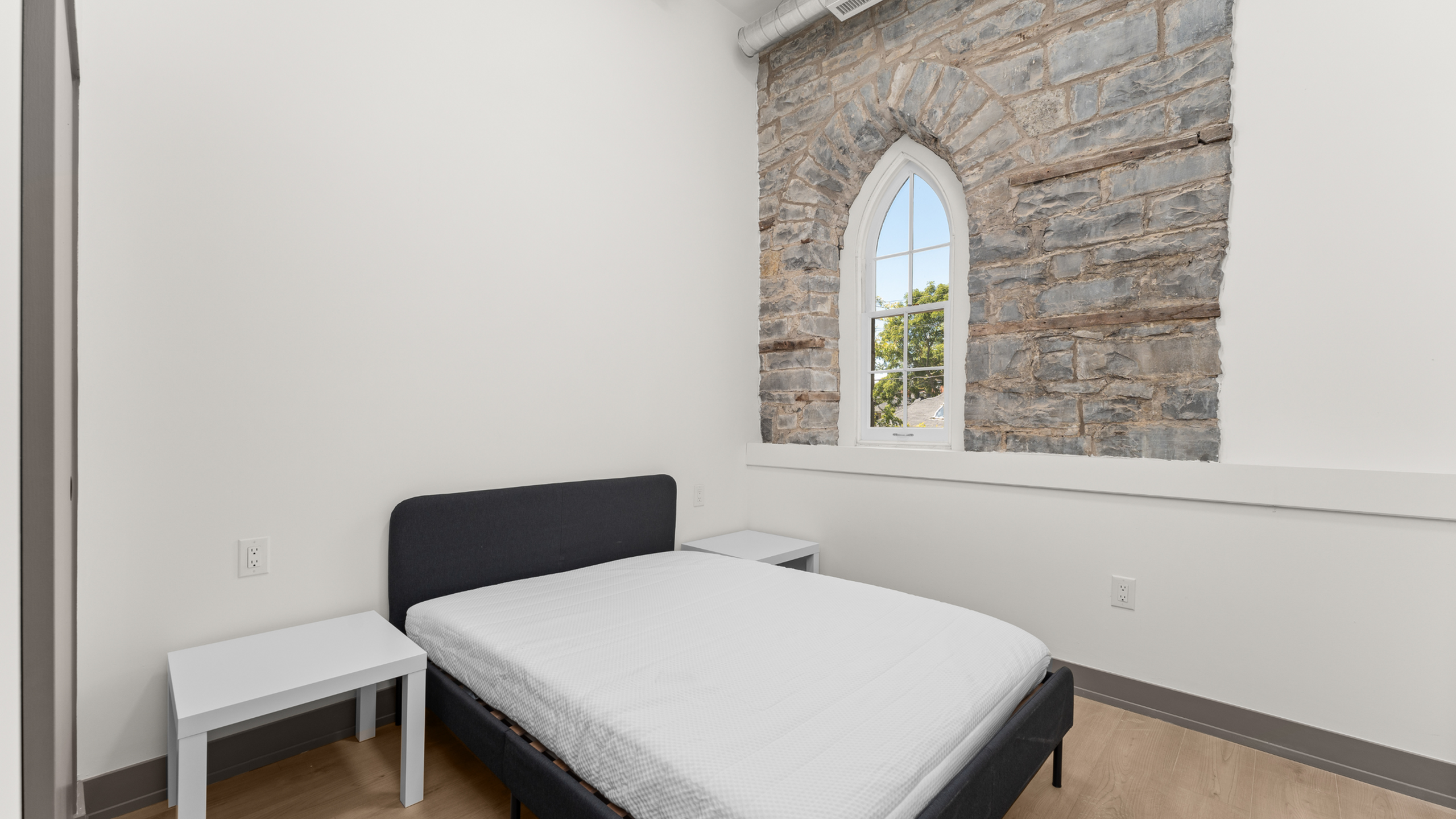 Charming bedroom with rustic stone wall and arched window, featuring a modern bed and sleek side tables.