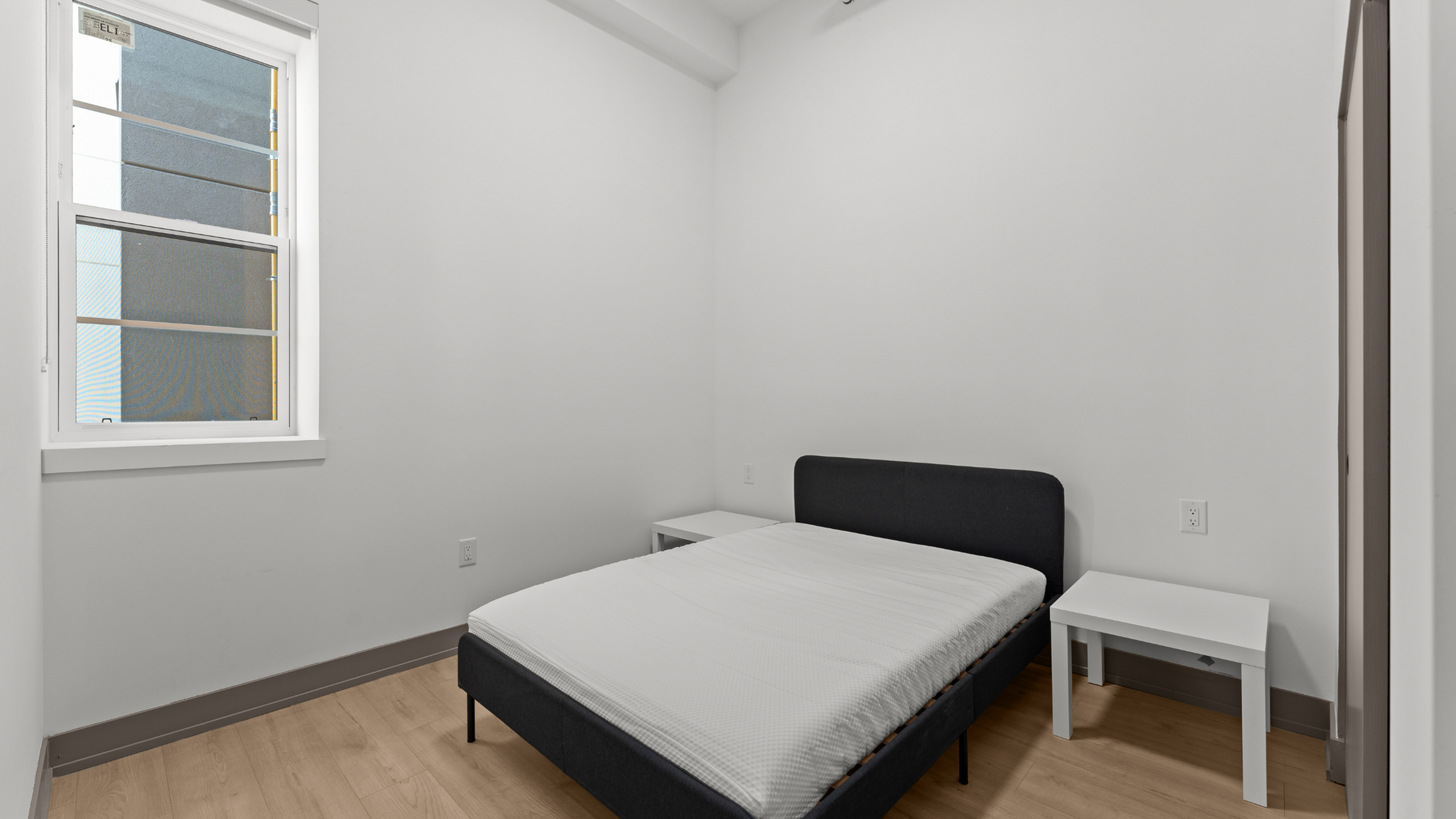 Cozy bedroom with minimalist design featuring a comfortable bed, wooden flooring, and ample natural light from the window.