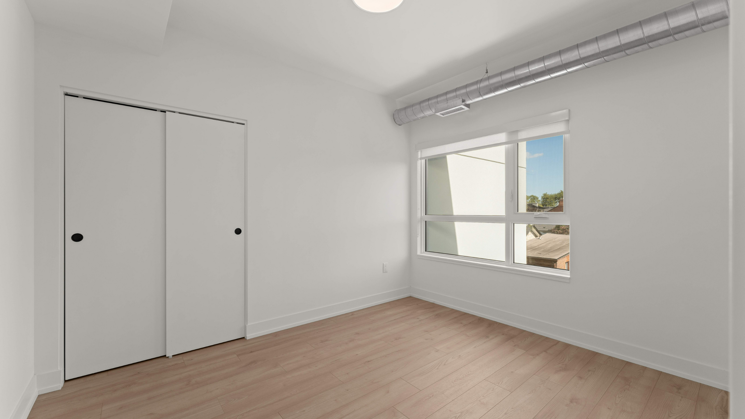 Bright modern bedroom with sleek closet doors, large window providing natural light, and stylish wood flooring.