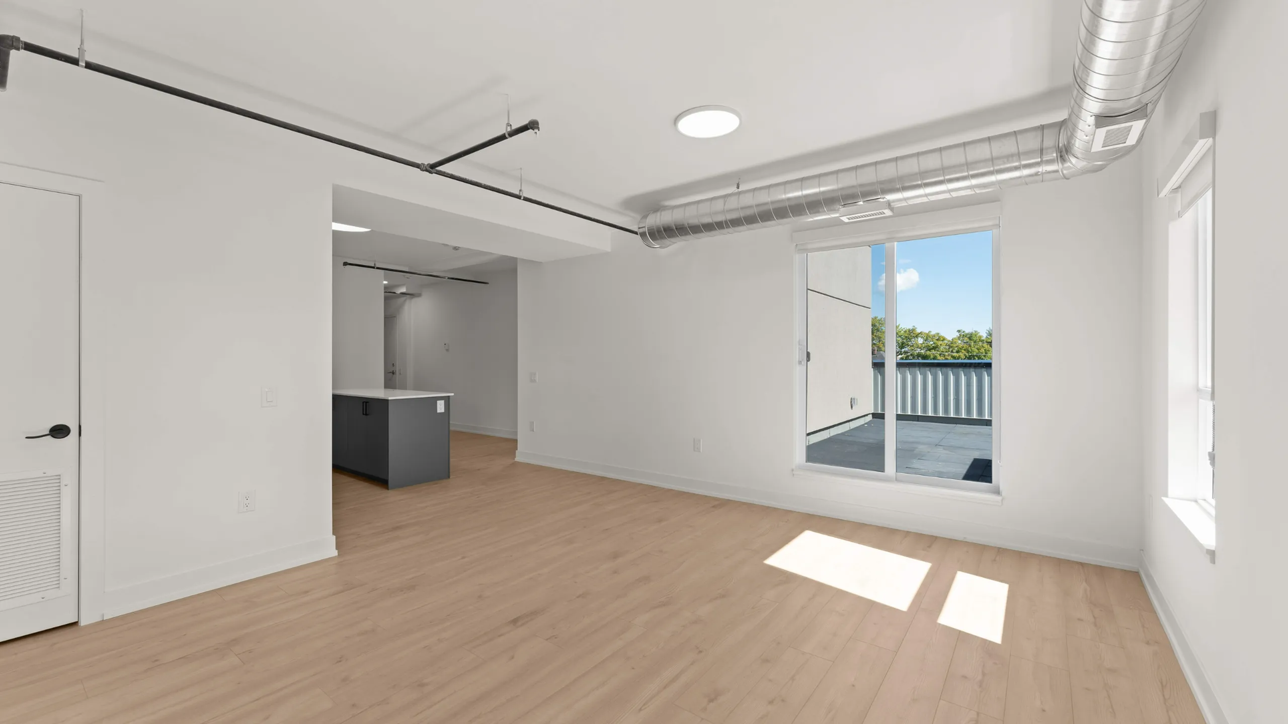 Modern apartment interior with open-concept layout featuring light wood flooring, large windows with outdoor terrace view, and exposed industrial ductwork.