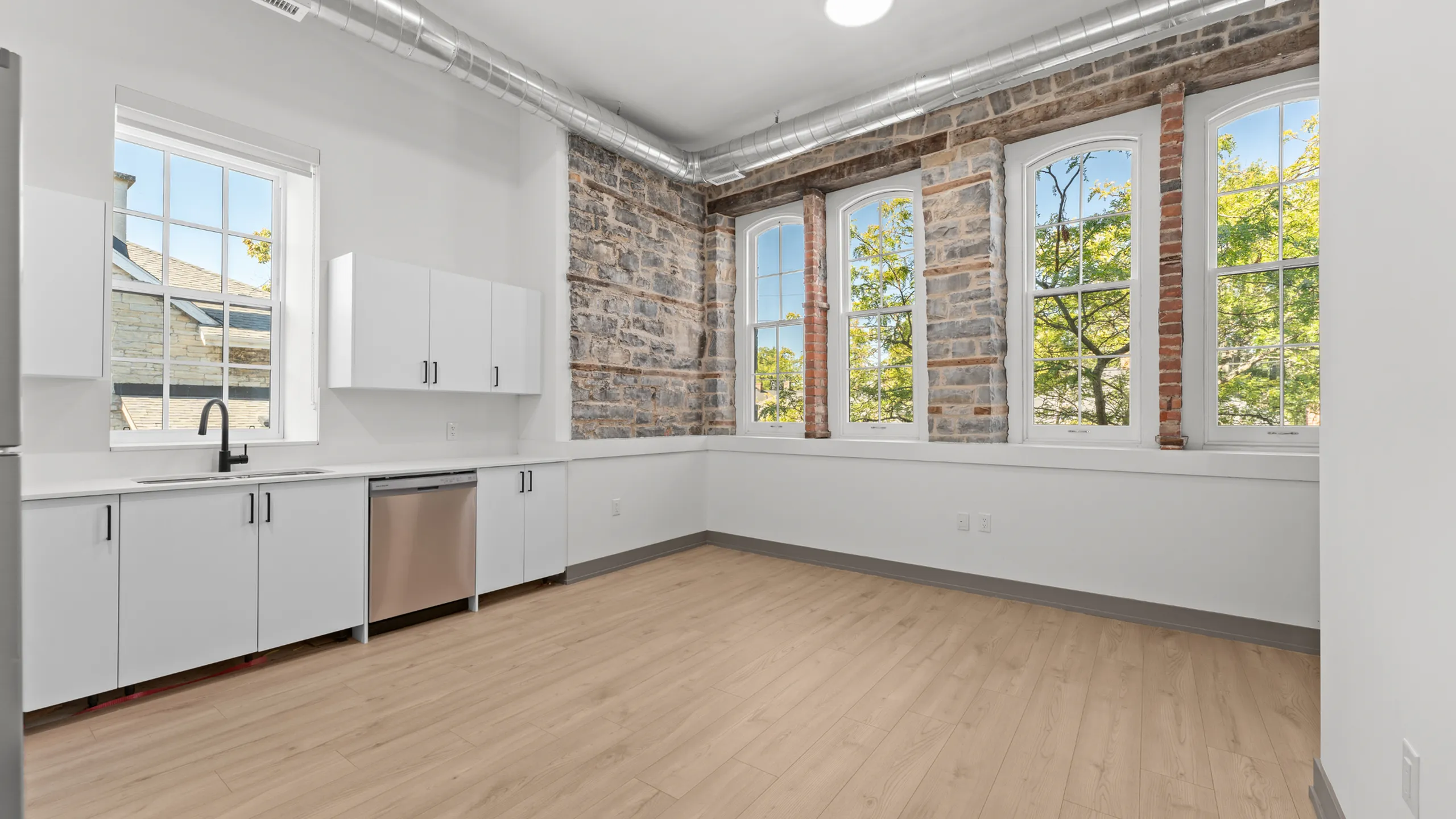 Bright industrial kitchen with exposed brick walls, large windows, and sleek white cabinets in a modern loft-style apartment.