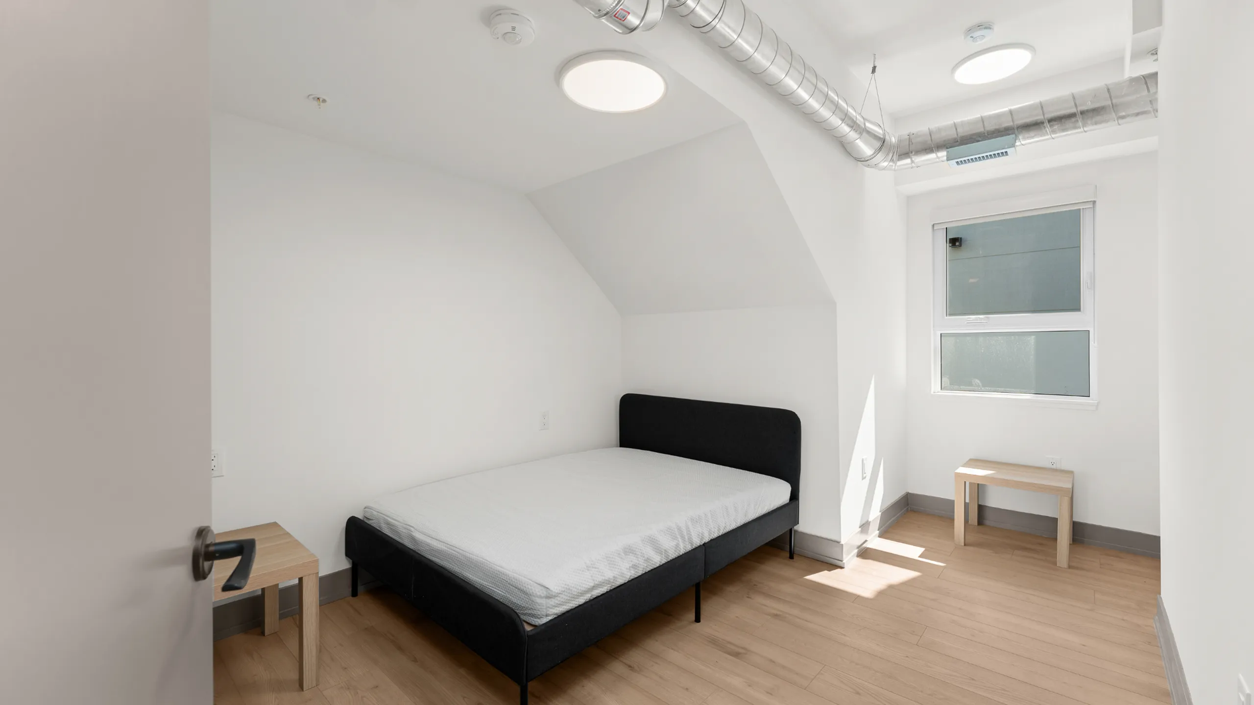 Bright and cozy bedroom with minimalist furnishings, featuring a comfortable bed, wooden flooring, and ample natural light from a large window.