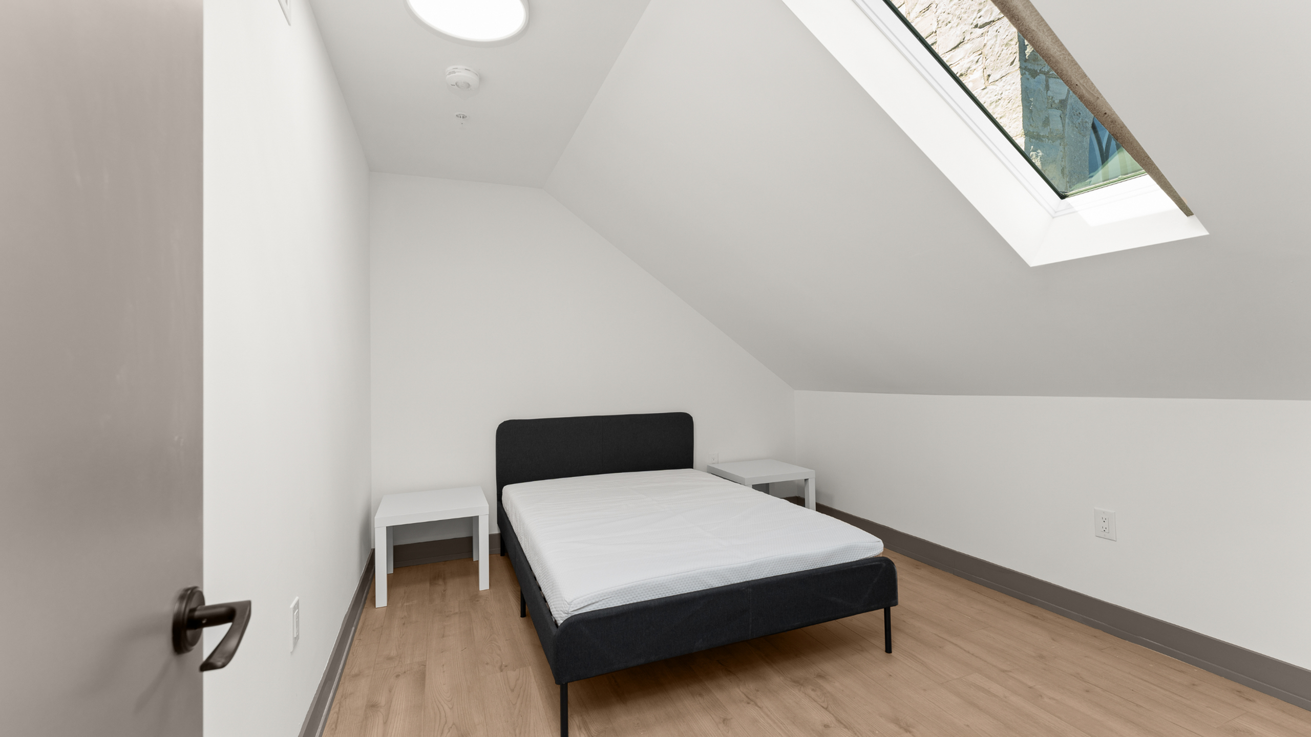 Bright minimalistic bedroom with a sloped ceiling and skylight, featuring a cozy bed and modern side tables on sleek wooden flooring.