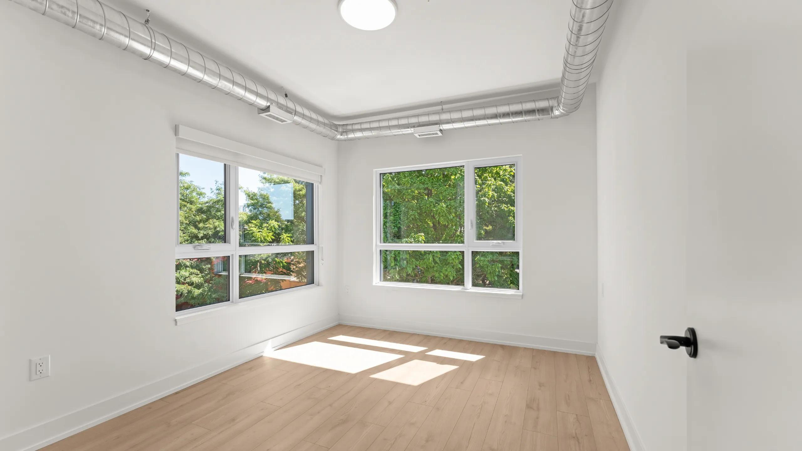 Bright and airy room featuring sleek wooden floors, large windows with lush green views, and modern exposed ductwork for an industrial touch.