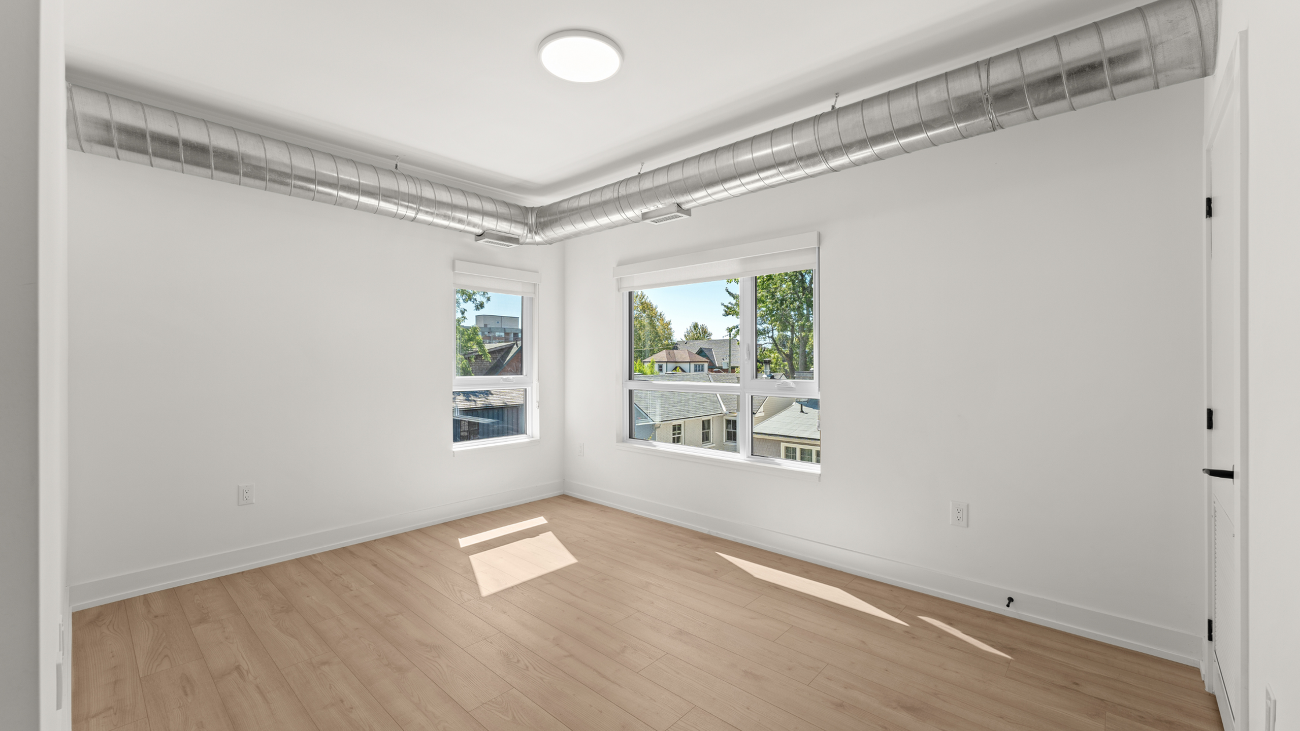 Bright, sunlit room with sleek hardwood floors and modern exposed ductwork, featuring large windows offering a scenic neighborhood view.