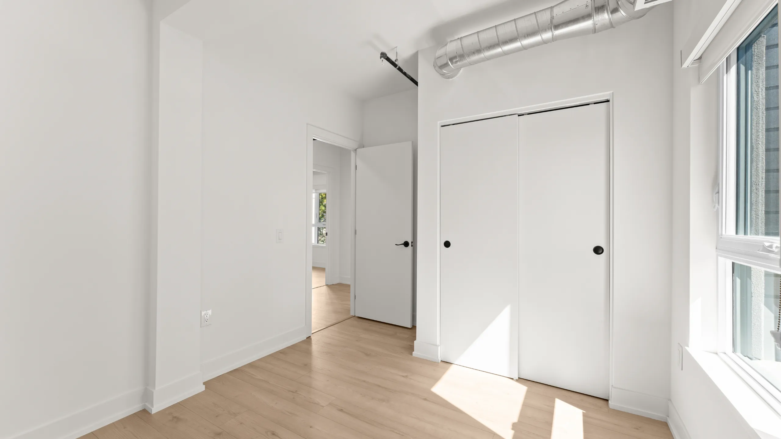 Bright room with large window, sleek wooden flooring, and modern closet doors. Abundant natural light creates an inviting atmosphere.