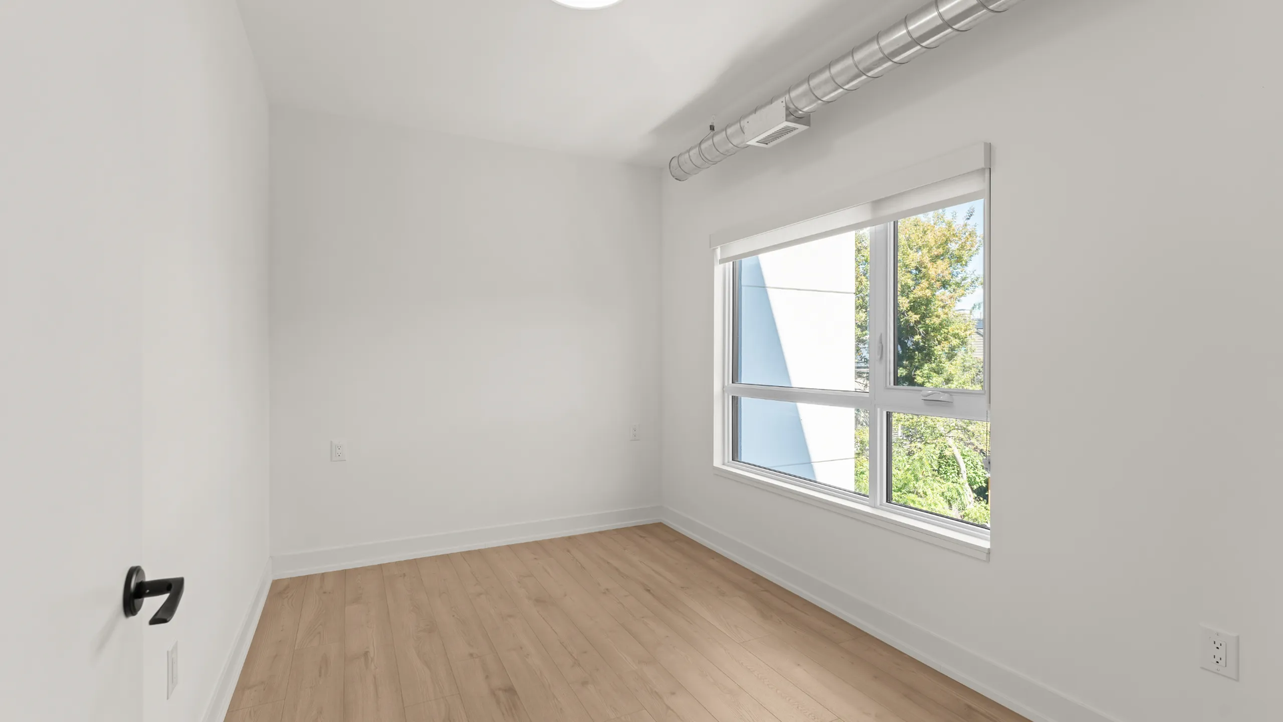 Bright room with large window and natural light, featuring sleek hardwood flooring and modern ductwork for a contemporary feel.