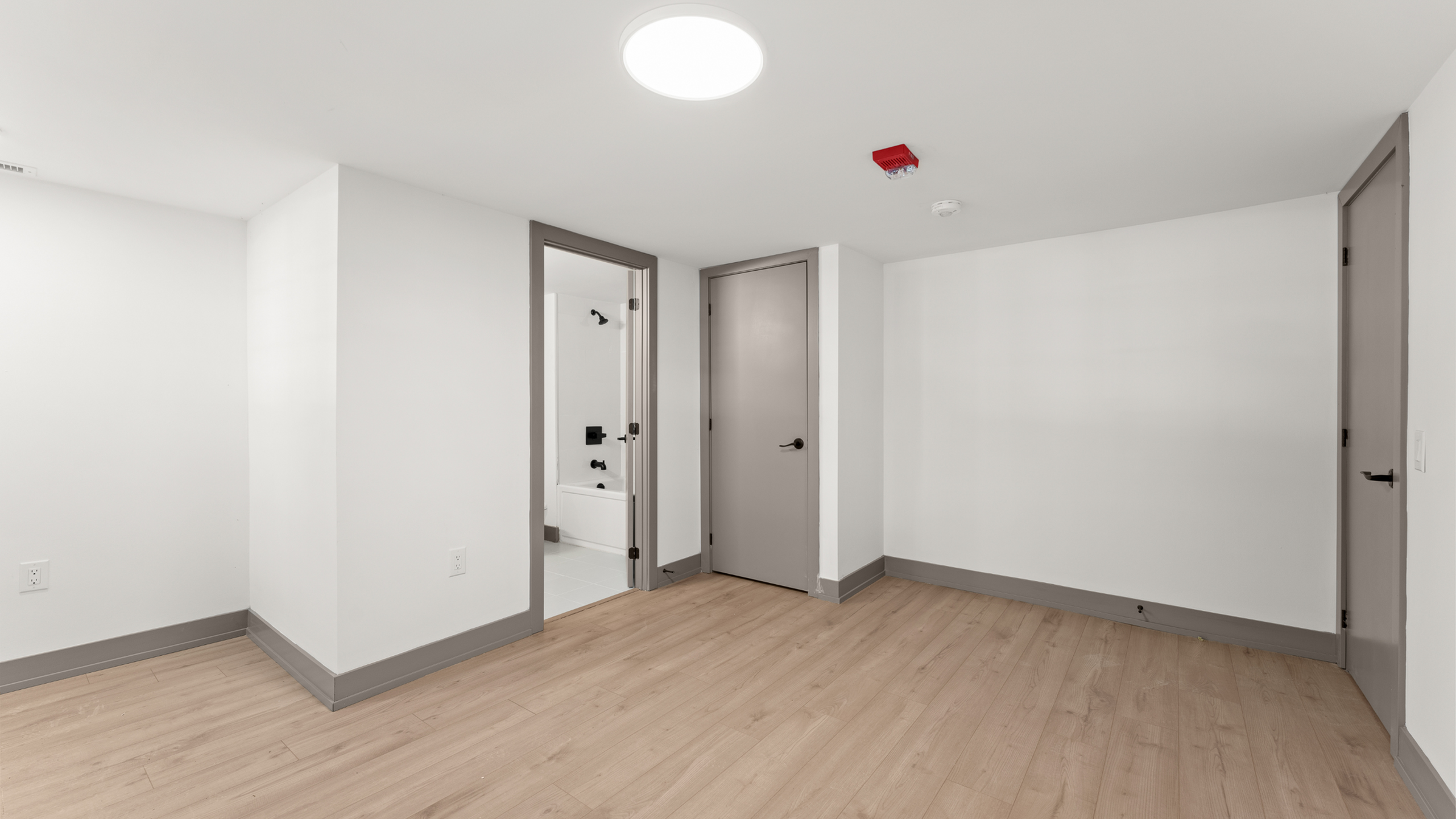 Bright, modern apartment room with sleek wooden floors and elegant white walls, featuring a view into a stylish bathroom with black fixtures.