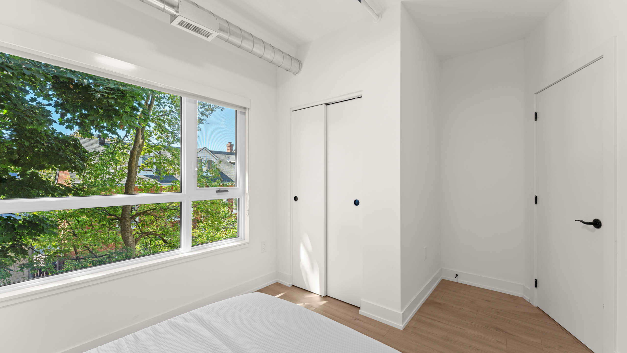 Bright bedroom with large window offering a scenic view of lush greenery, featuring sleek white closet and modern hardwood flooring.