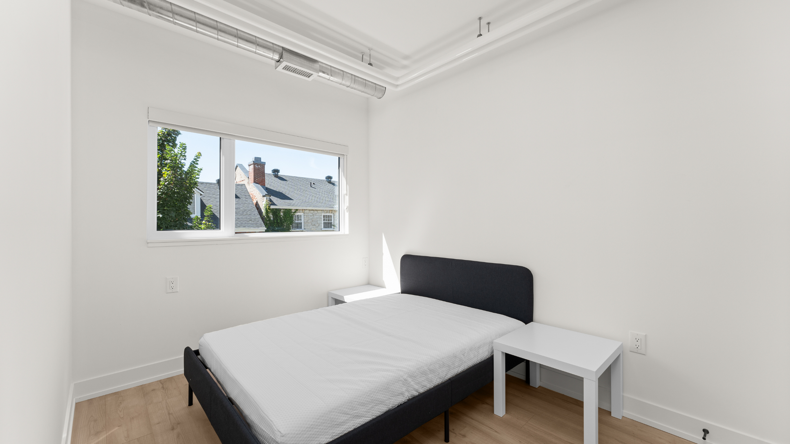 Bright and modern bedroom with a minimalist design, featuring a comfortable bed, sleek side tables, and natural light streaming in through a window with a cityscape view.