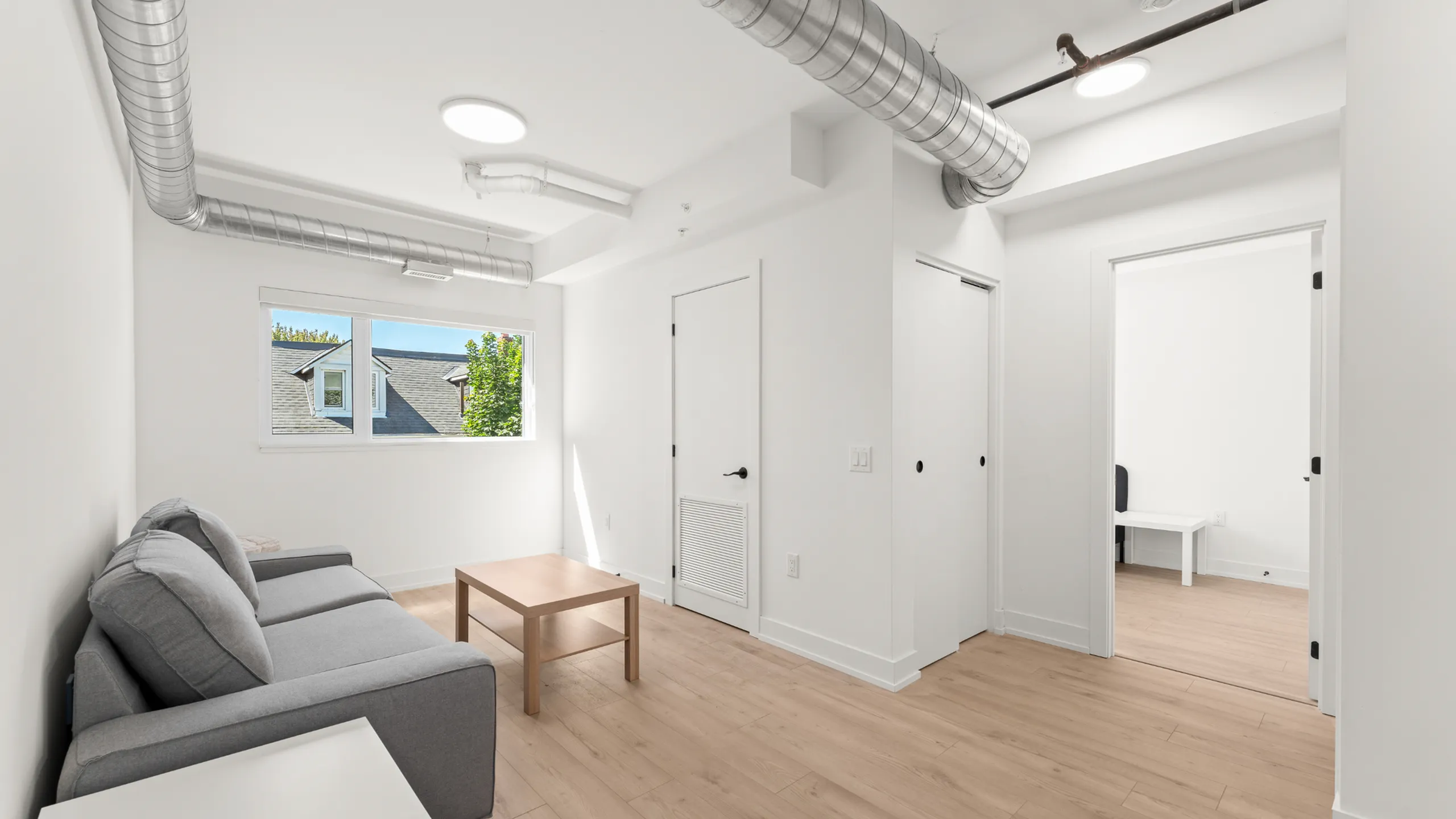 Modern apartment interior with sleek exposed ductwork, cozy gray sofa, and abundant natural light from a large window overlooking a charming neighborhood.