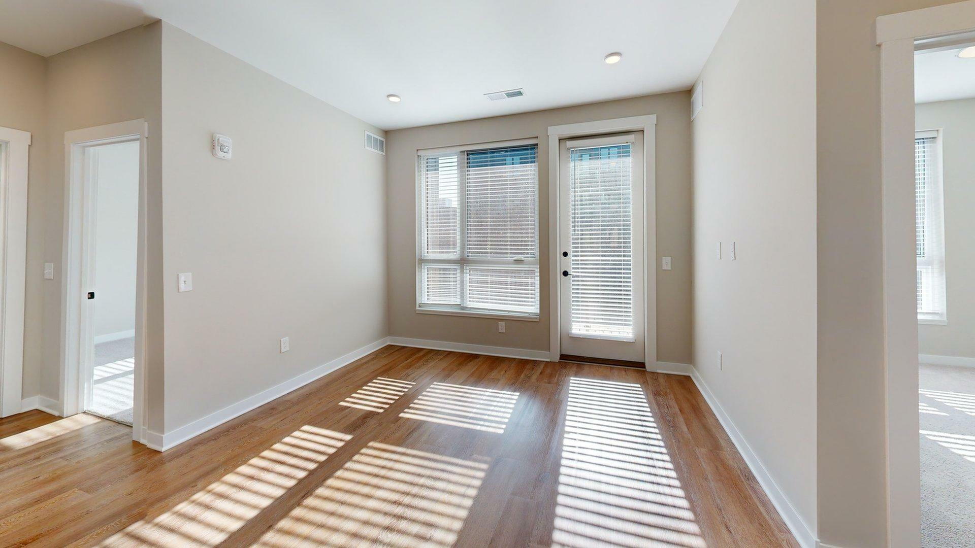 Bright interior view of a modern apartment, featuring large windows with blinds, hardwood floors, and white walls.