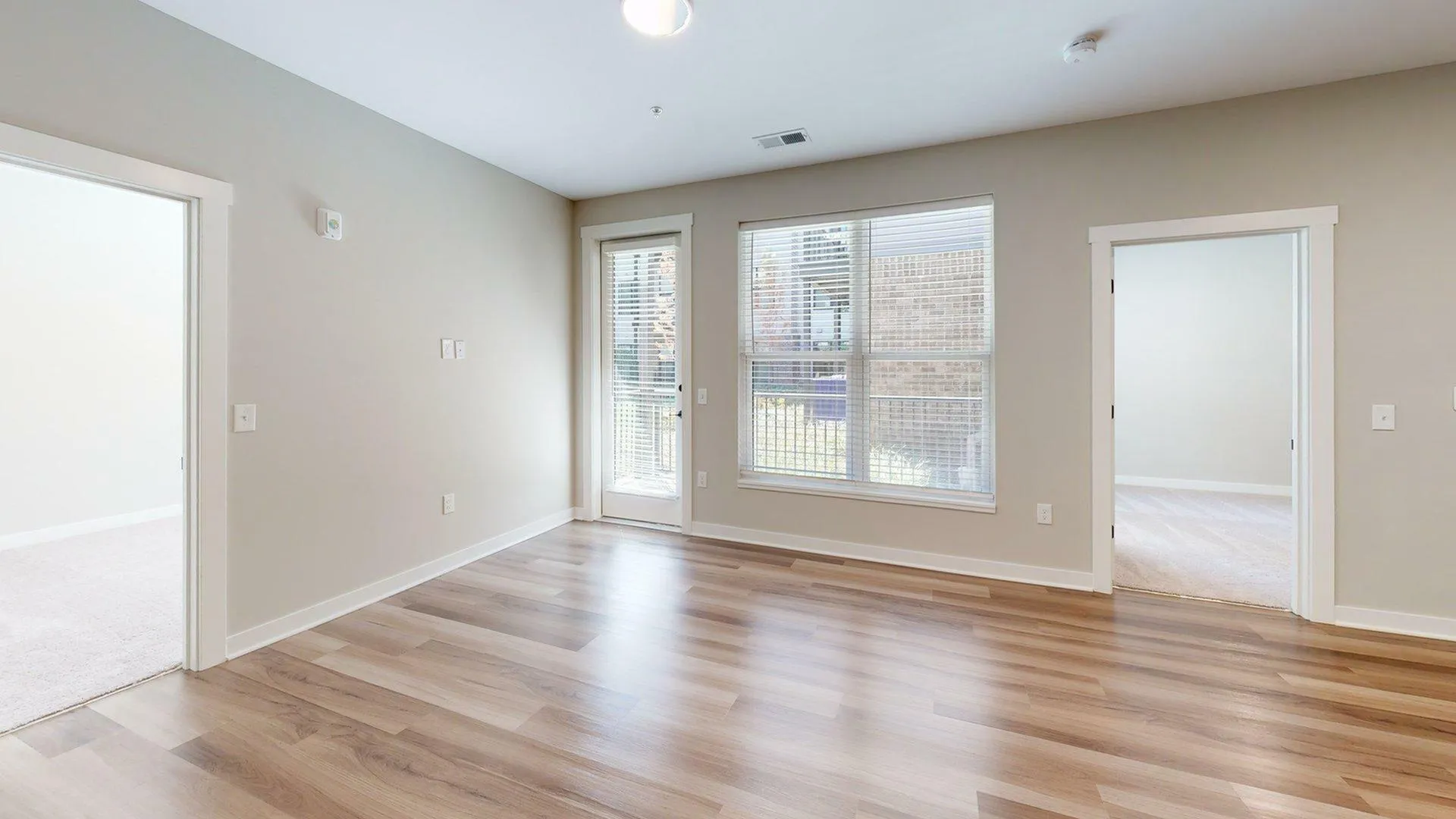 Bright modern living room with sleek hardwood floors, large windows letting in natural light, and access to adjacent rooms and exterior.
