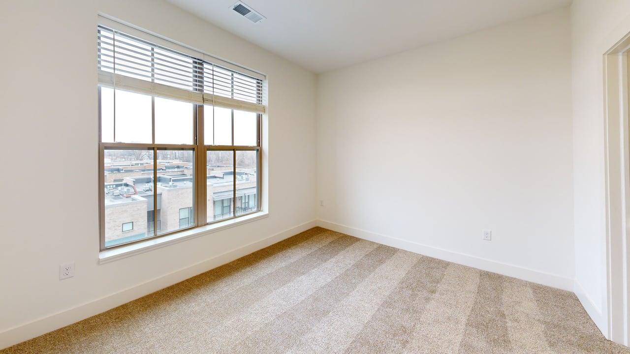 Bright and spacious room with large window offering city views, featuring neutral tones and plush carpet.