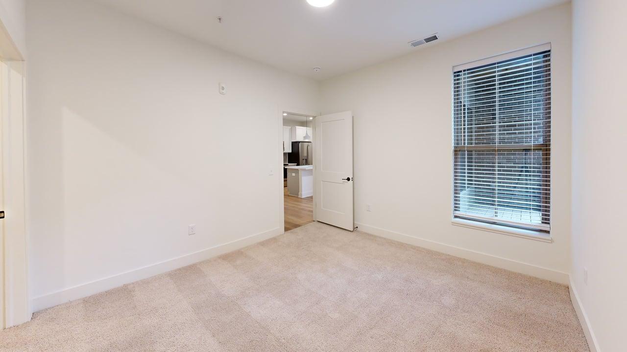 Bright bedroom with plush carpeting, large window, and view into a modern kitchen with sleek appliances.