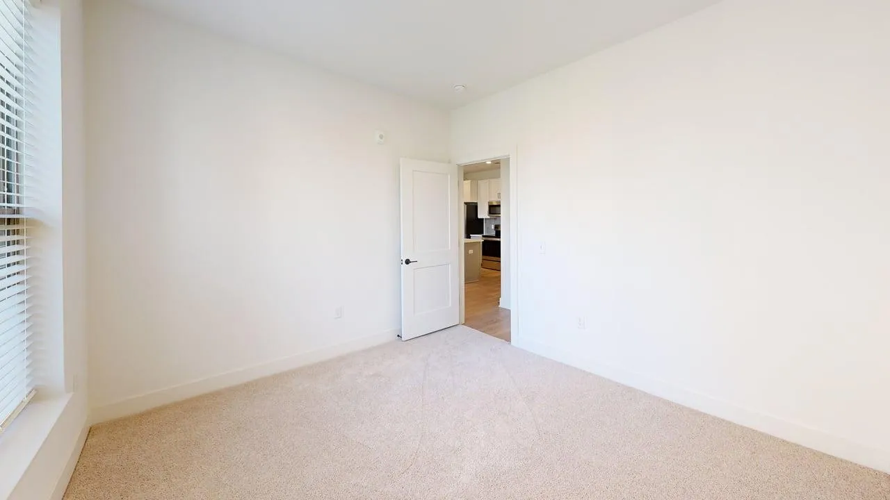 Bright and airy bedroom with plush carpet and a door leading to a modern kitchen; ideal for comfort and convenience.