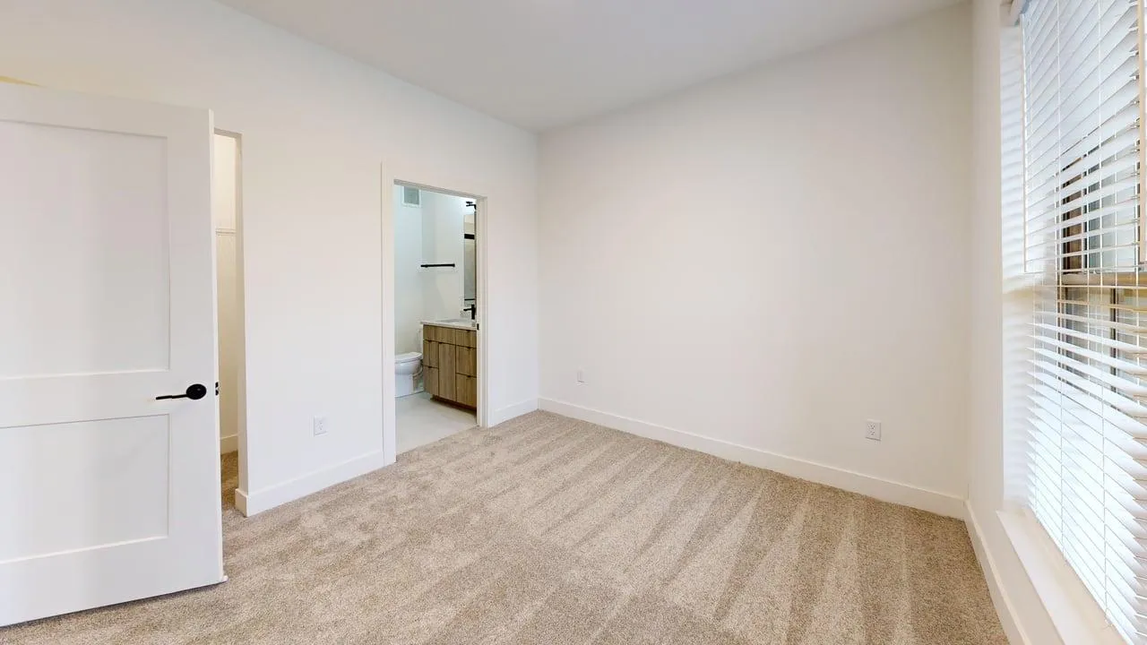Bright and cozy bedroom with carpet flooring, featuring an en-suite bathroom and large window with blinds.