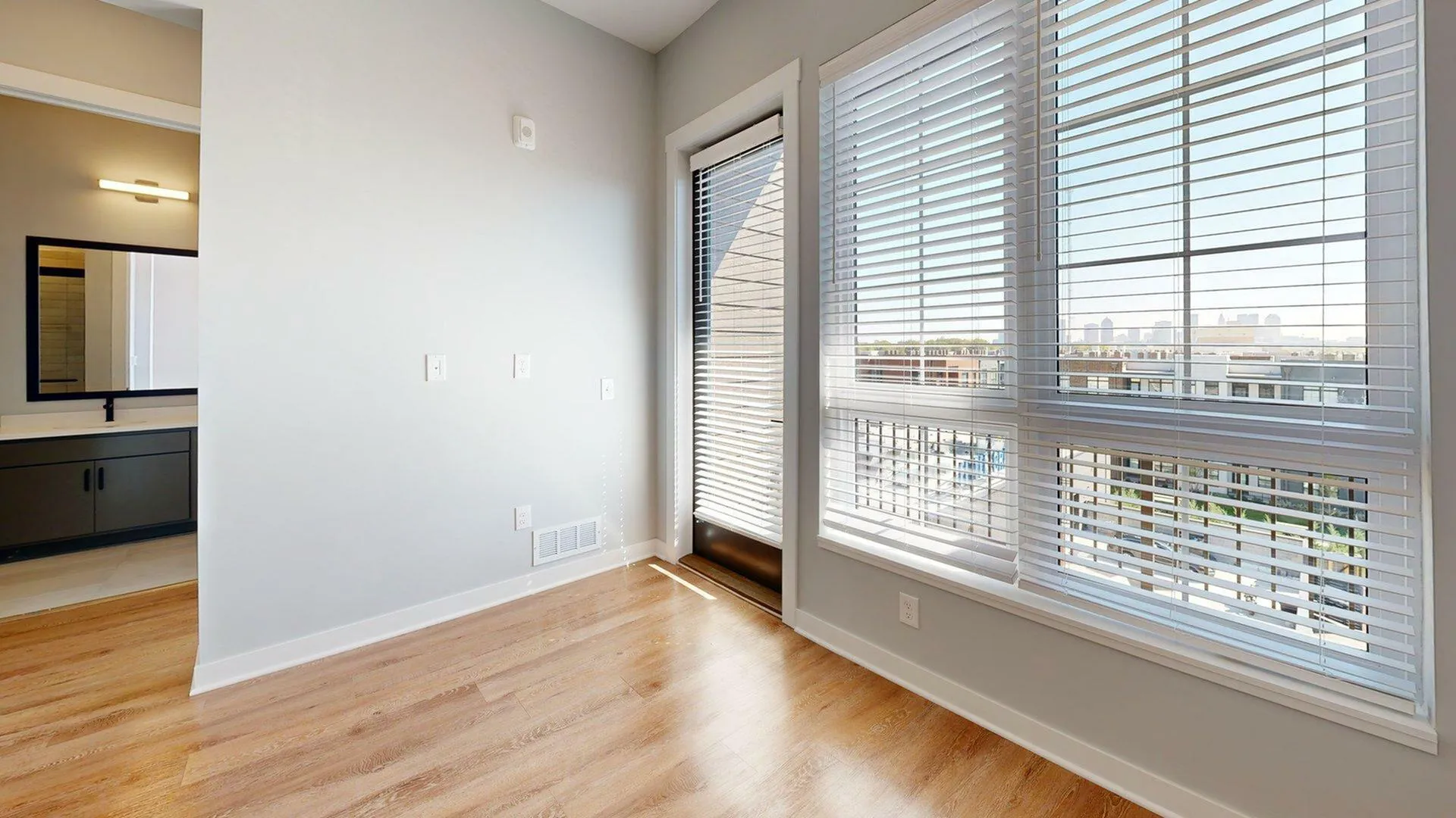 Bright apartment interior with large windows and modern wood flooring, offering a view of the city skyline.