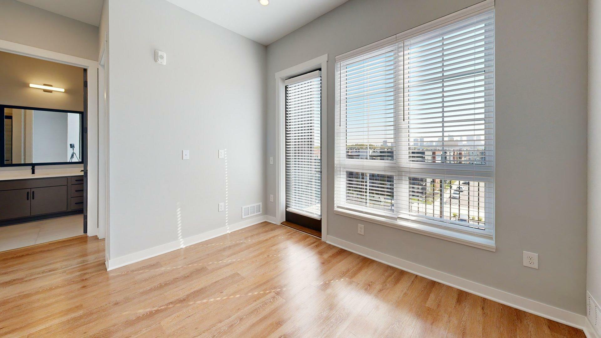Sunlit room with modern wood flooring and large windows offering city views, adjacent to a stylish bathroom.