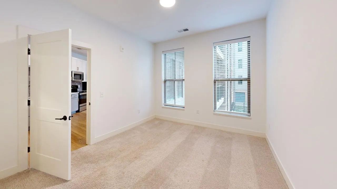 Bright bedroom with large windows, plush carpeting, and a view into a modern kitchen. Ideal for comfortable living.