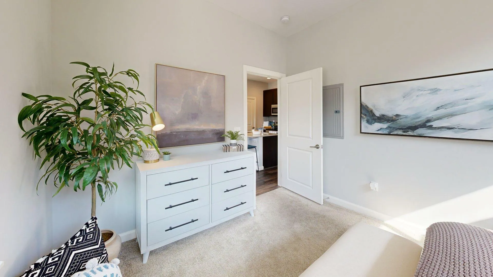 Bright and airy bedroom with modern decor, featuring a chic dresser, vibrant indoor plant, and elegant wall art, leading to a stylish kitchen.