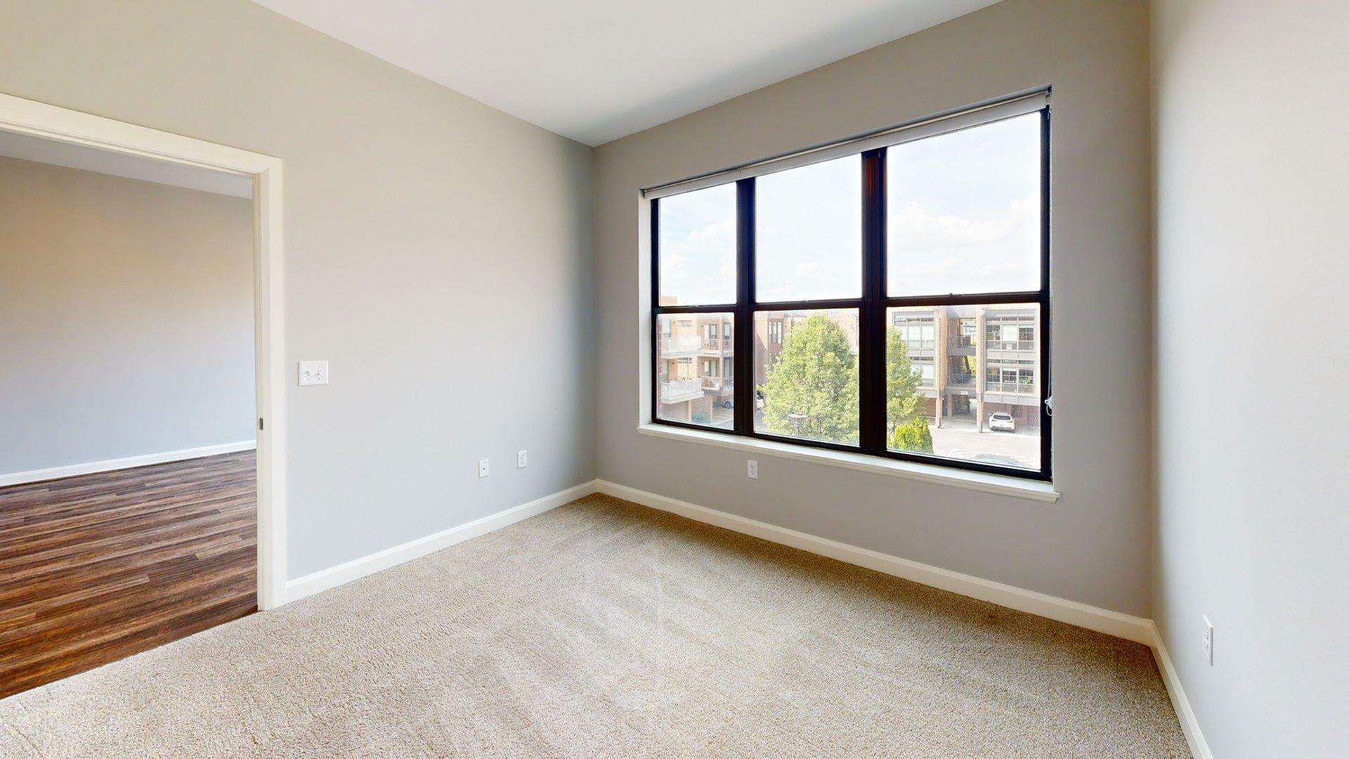 Bright and inviting room with large window providing ample natural light, plush carpeting, and a view of modern residences.