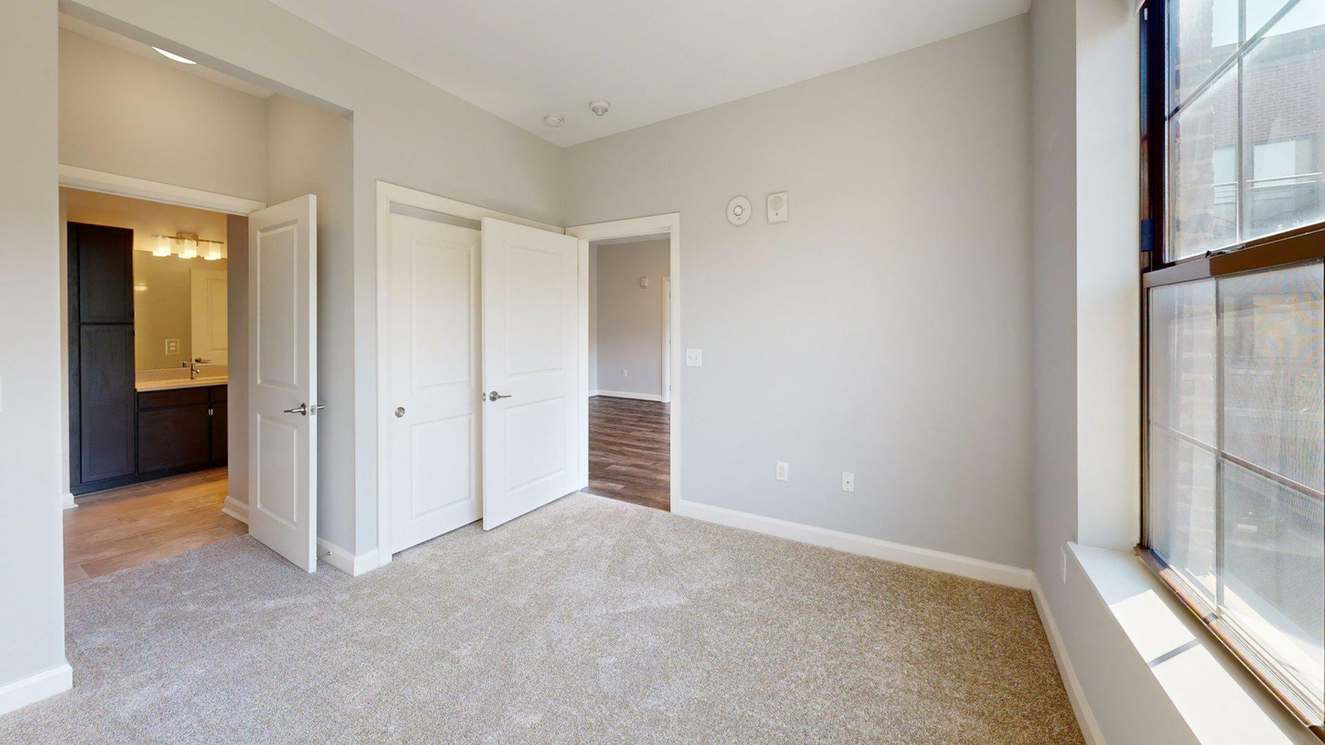 Bright and inviting bedroom with plush carpet, large window, and access to a modern bathroom through double doors.