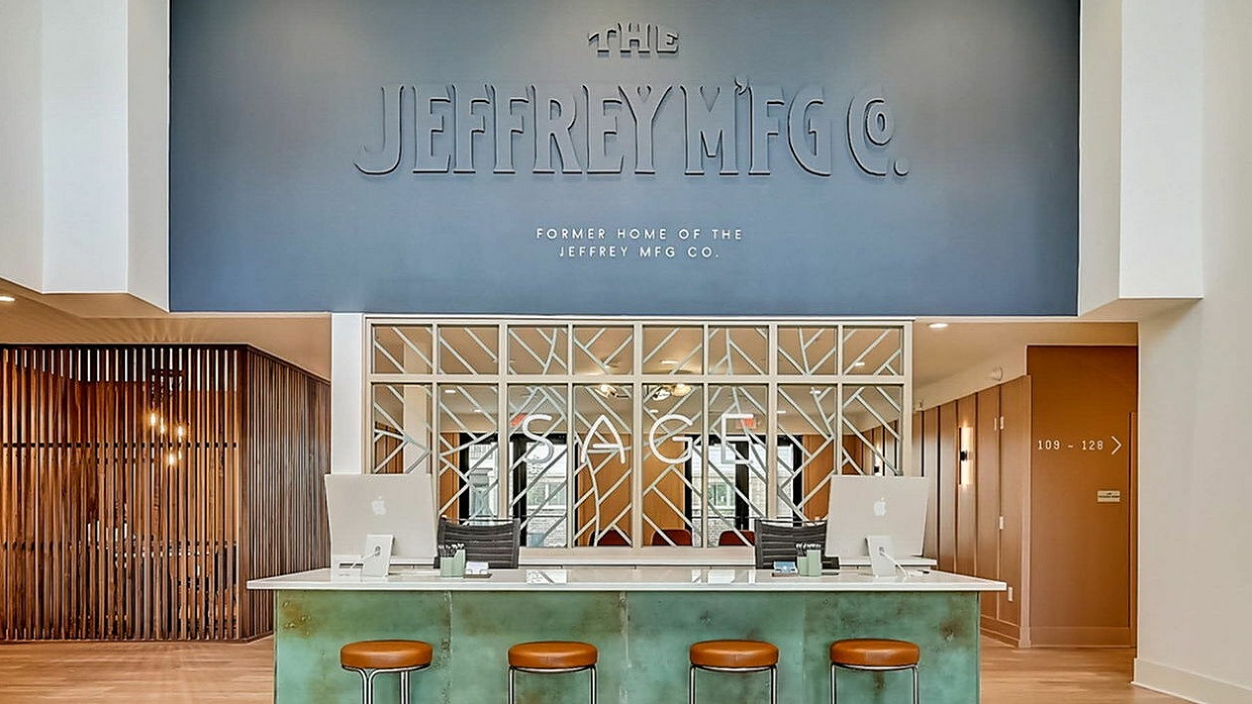 Modern apartment lobby featuring a sleek reception desk with stylish stools, warm wooden accents, and a grand sign of The Jeffrey Mfg Co.