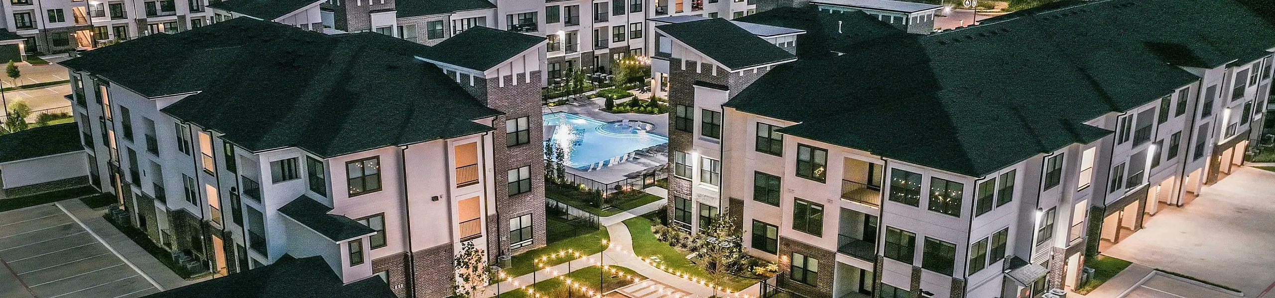 Elegant apartment complex at dusk with beautifully lit pathways and an inviting central pool area, surrounded by modern three-story buildings.