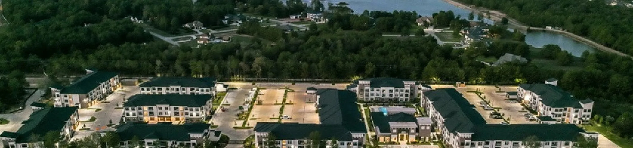 Scenic aerial view of a modern apartment complex surrounded by lush greenery and a serene lake, offering tranquil living near nature.