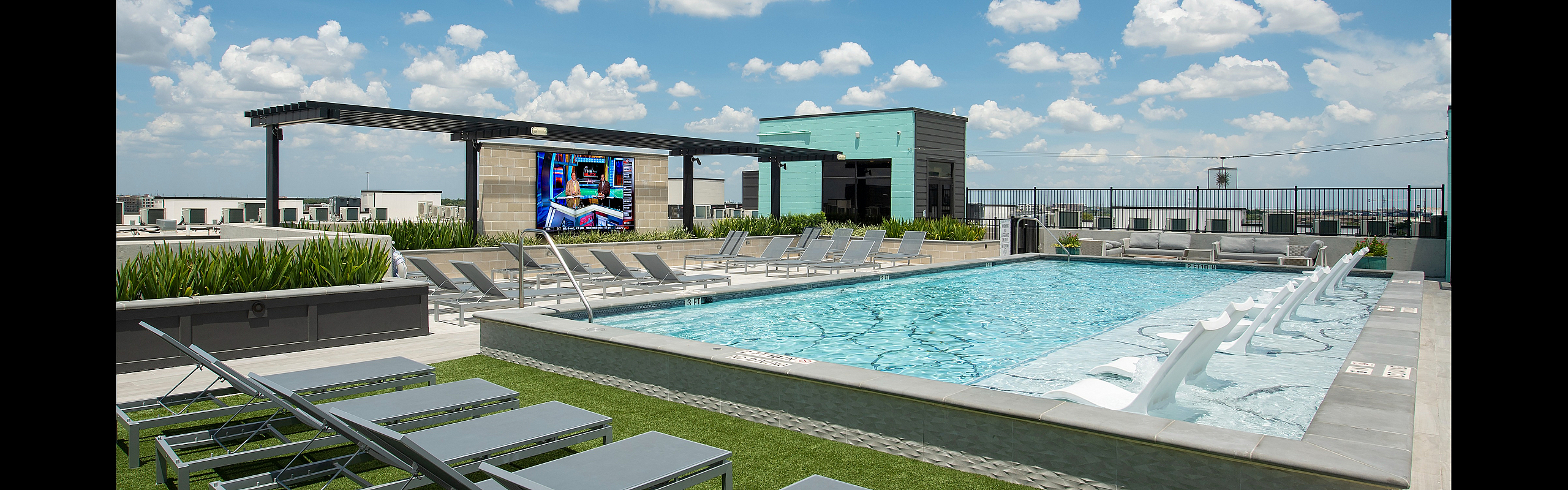 Luxurious rooftop pool with stylish lounge chairs, vibrant greenery, and a large TV screen under a blue sky.