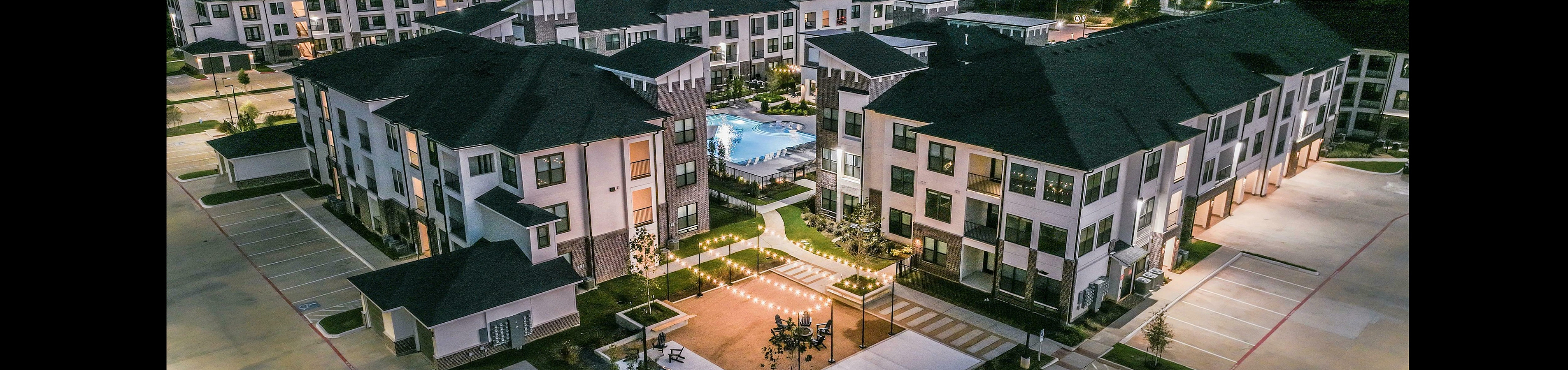Modern apartment complex at dusk featuring illuminated pathways, a central courtyard with a swimming pool, and ample parking spaces.