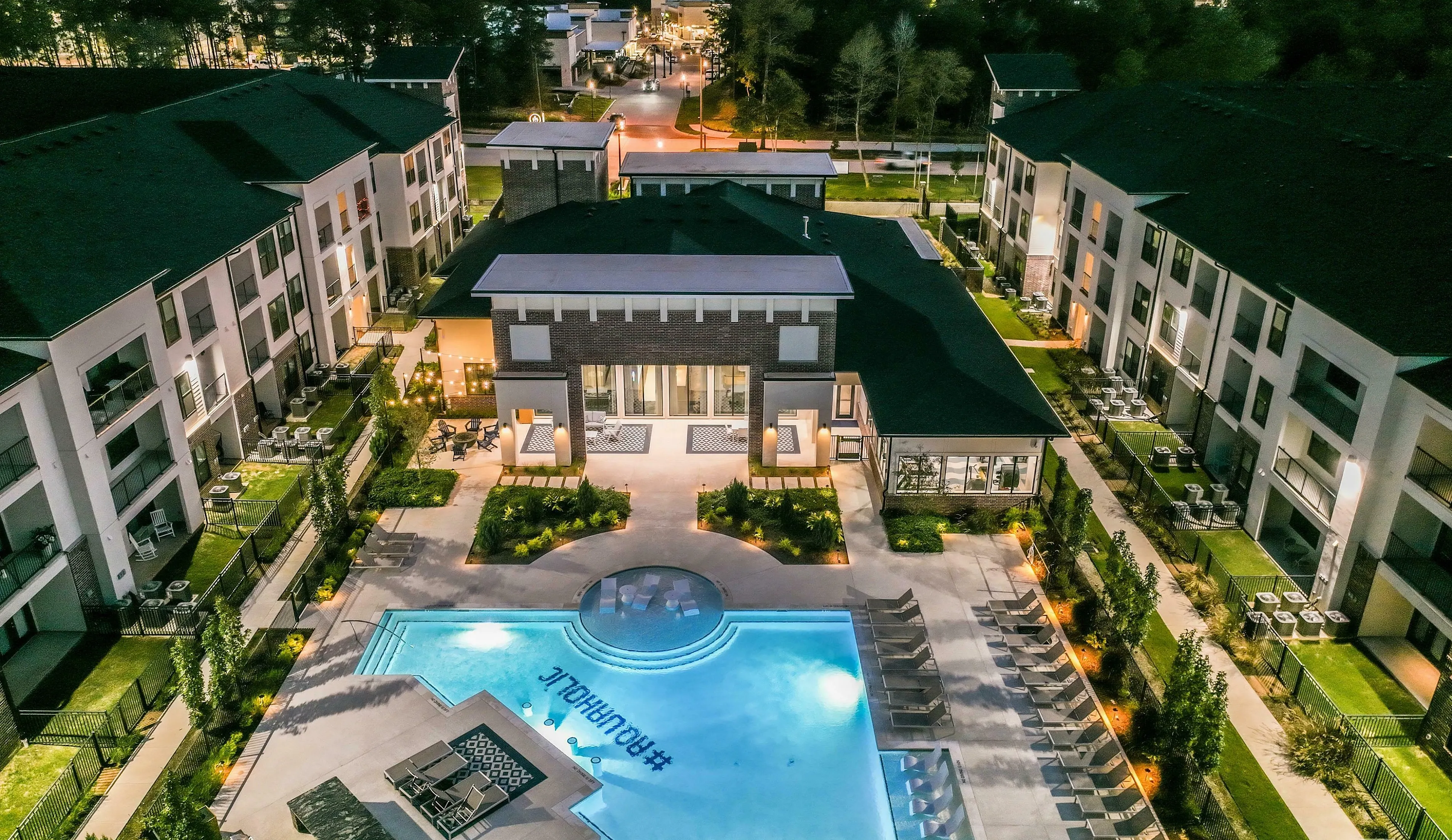 Luxurious apartment complex at dusk featuring a stunning illuminated pool, surrounded by elegant modern buildings and lush landscaped gardens.