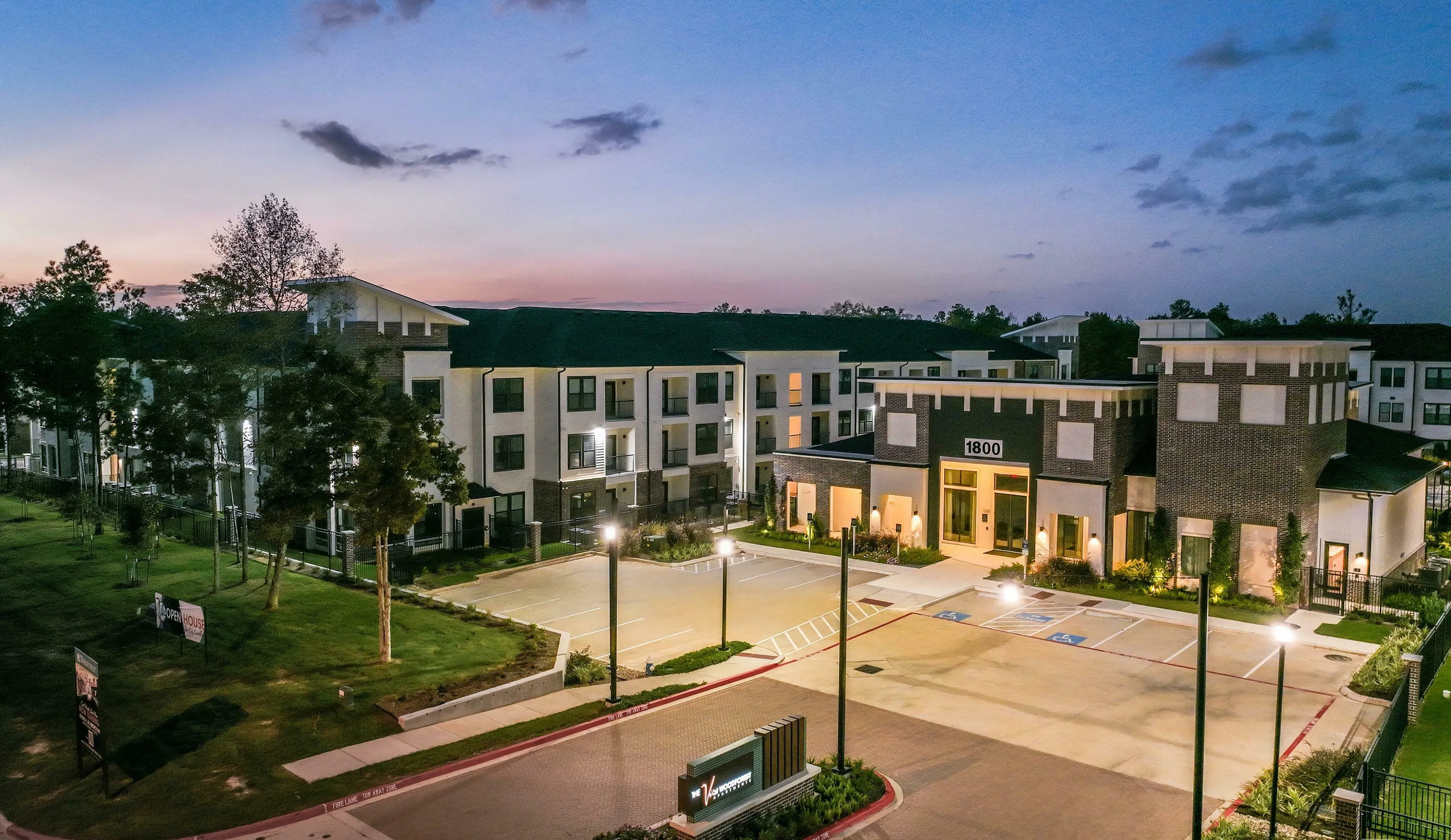 Scenic twilight view of a modern apartment complex with elegant architecture, lush greenery, and paved paths, surrounded by a serene backdrop of a colorful sky.