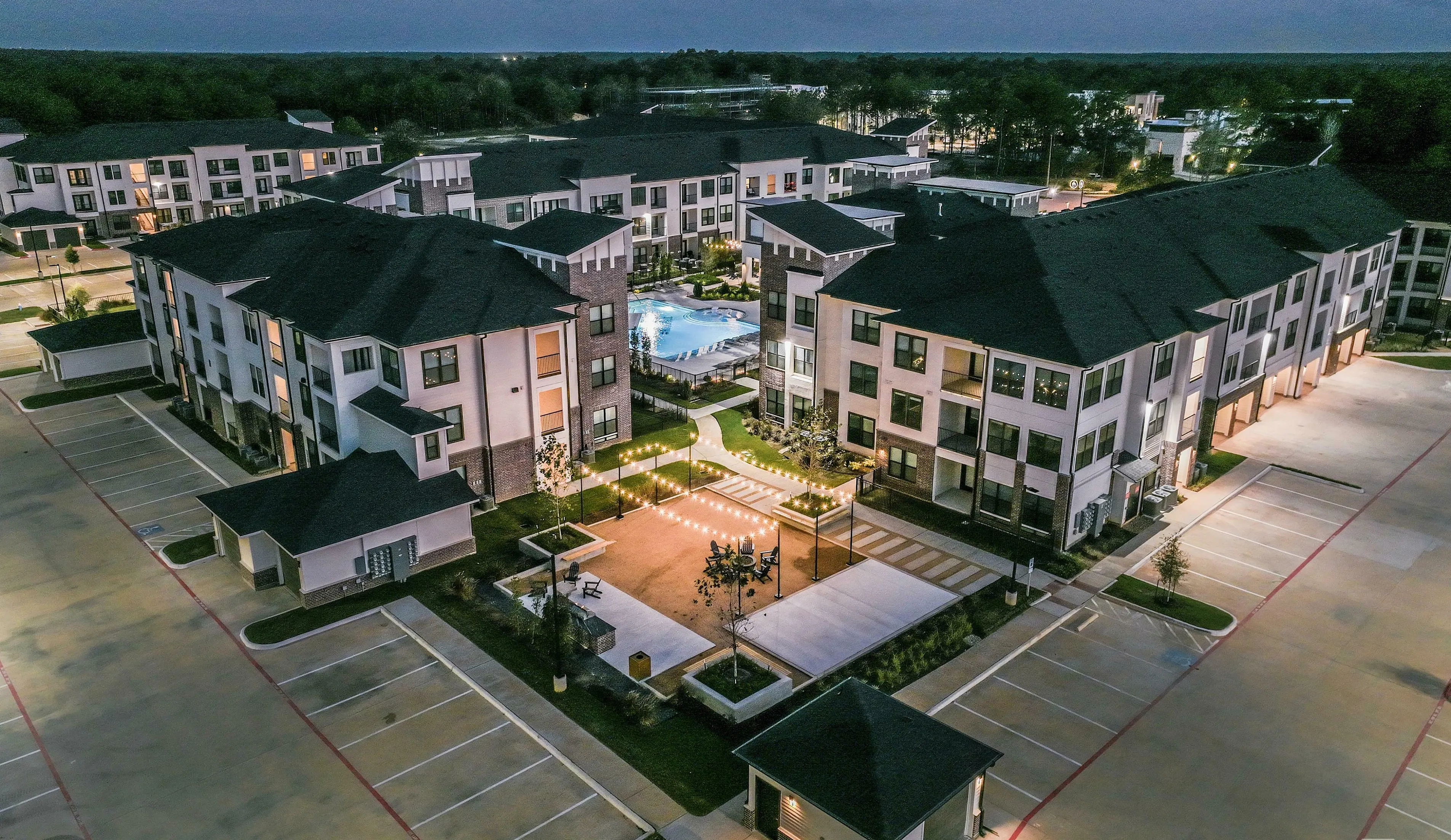 Elegant aerial view of a modern apartment complex at dusk, showcasing beautifully lit courtyards and a central swimming pool surrounded by contemporary buildings.