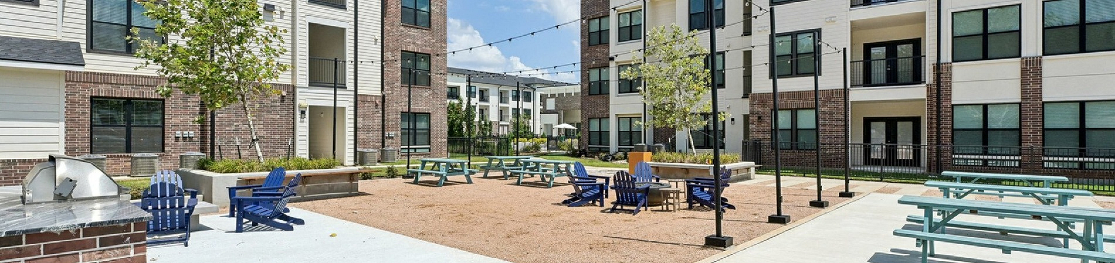 Modern apartment complex with spacious outdoor courtyard featuring picnic tables, seating areas, and vibrant greenery under a sunny sky.