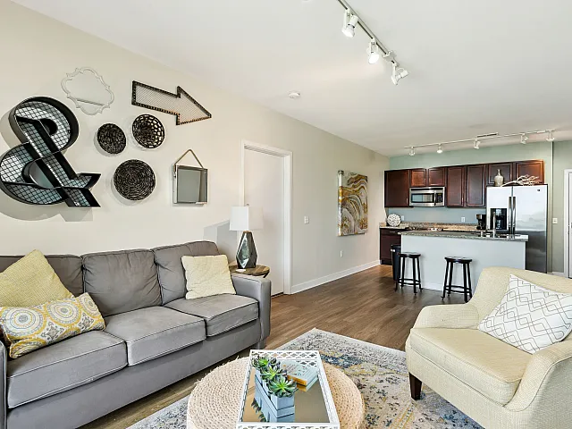 Modern open-plan living area with cozy seating, stylish decor on a neutral wall, and a sleek kitchen with dark cabinetry and stainless appliances.