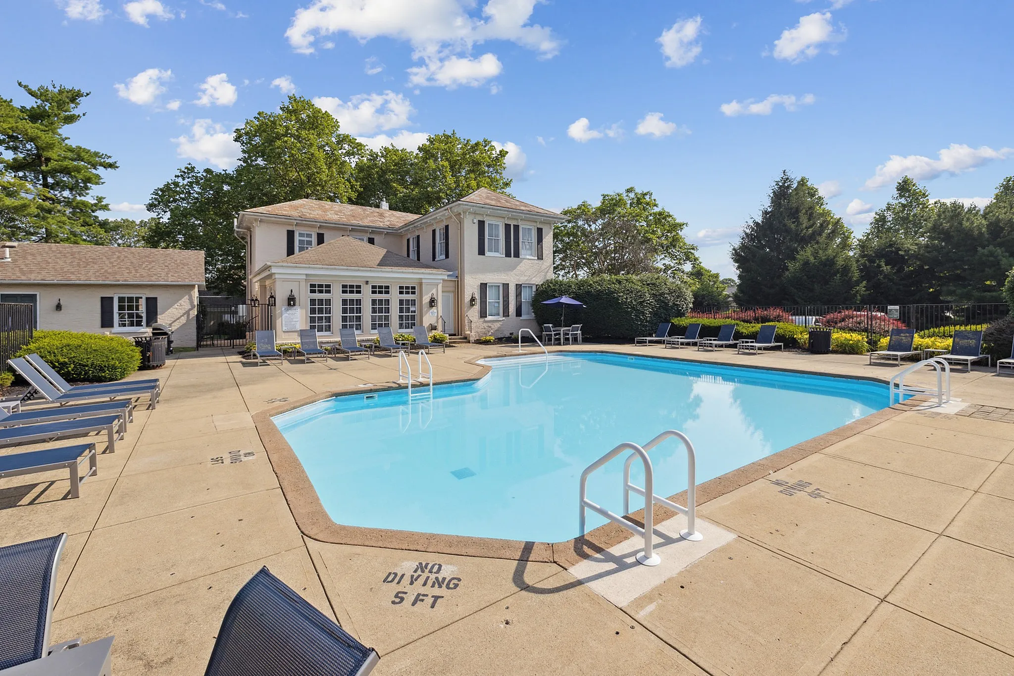 Inviting outdoor swimming pool area with lounge chairs, surrounded by lush greenery and a charming building under a bright blue sky.